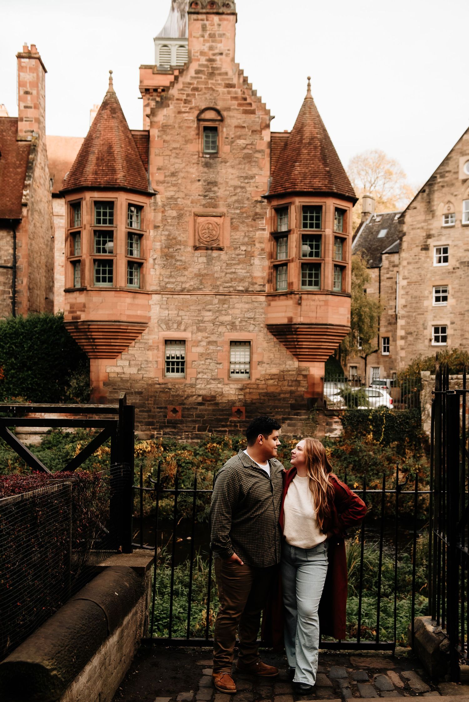 Couple shares intimate moment along stone wall with historic Scottish architecture in background.
