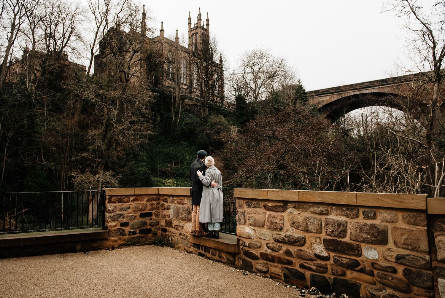 Stone bridge arches over a river with gothic architecture and church spires visible in the distance.