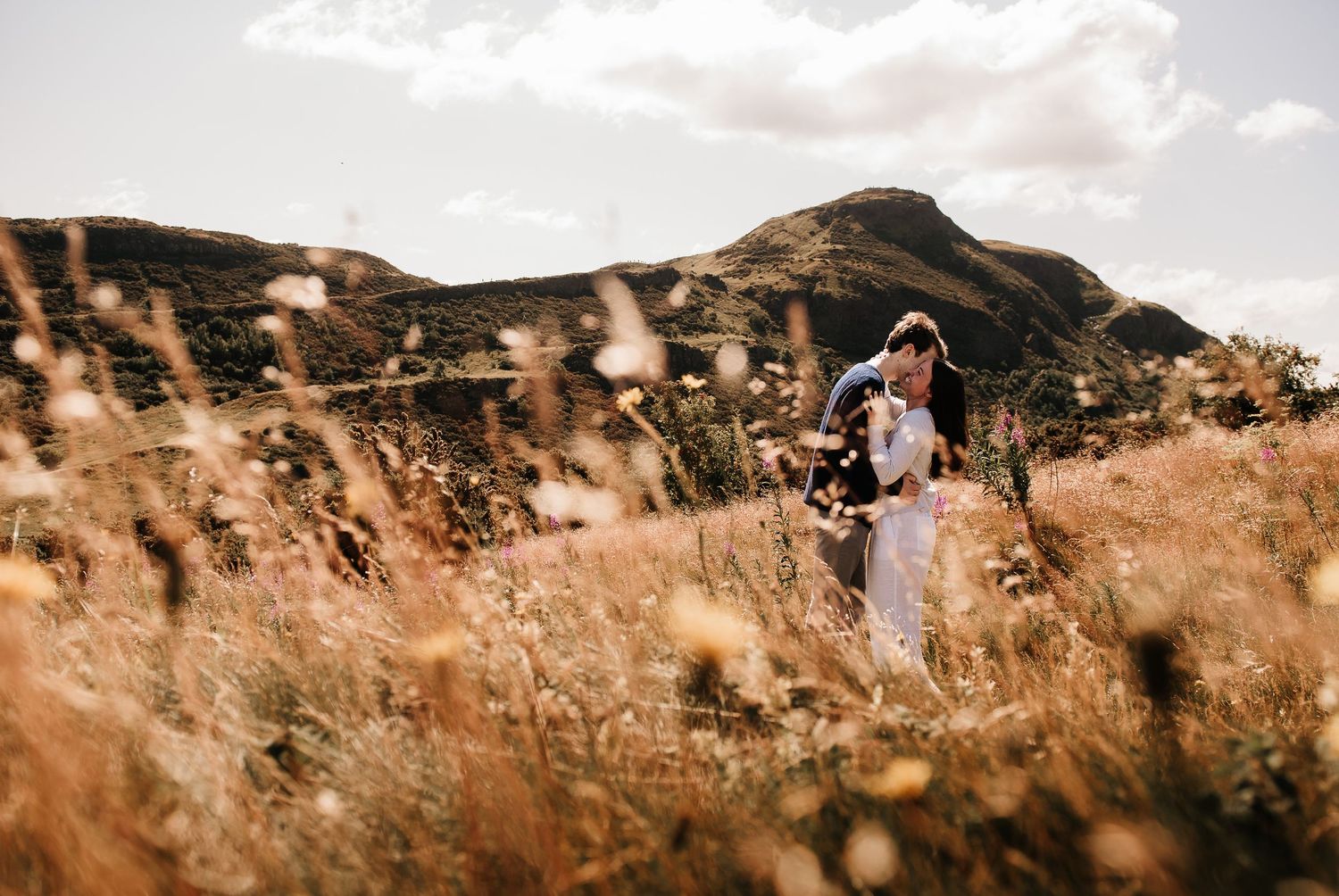 A panoramic view of a couple embracing on a mountainous hillside during golden hour.