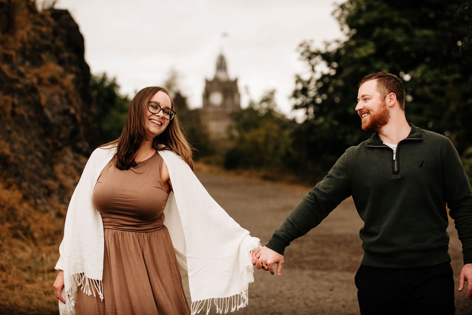 Couple walking hand in hand through an autumn landscape with historic buildings in view.