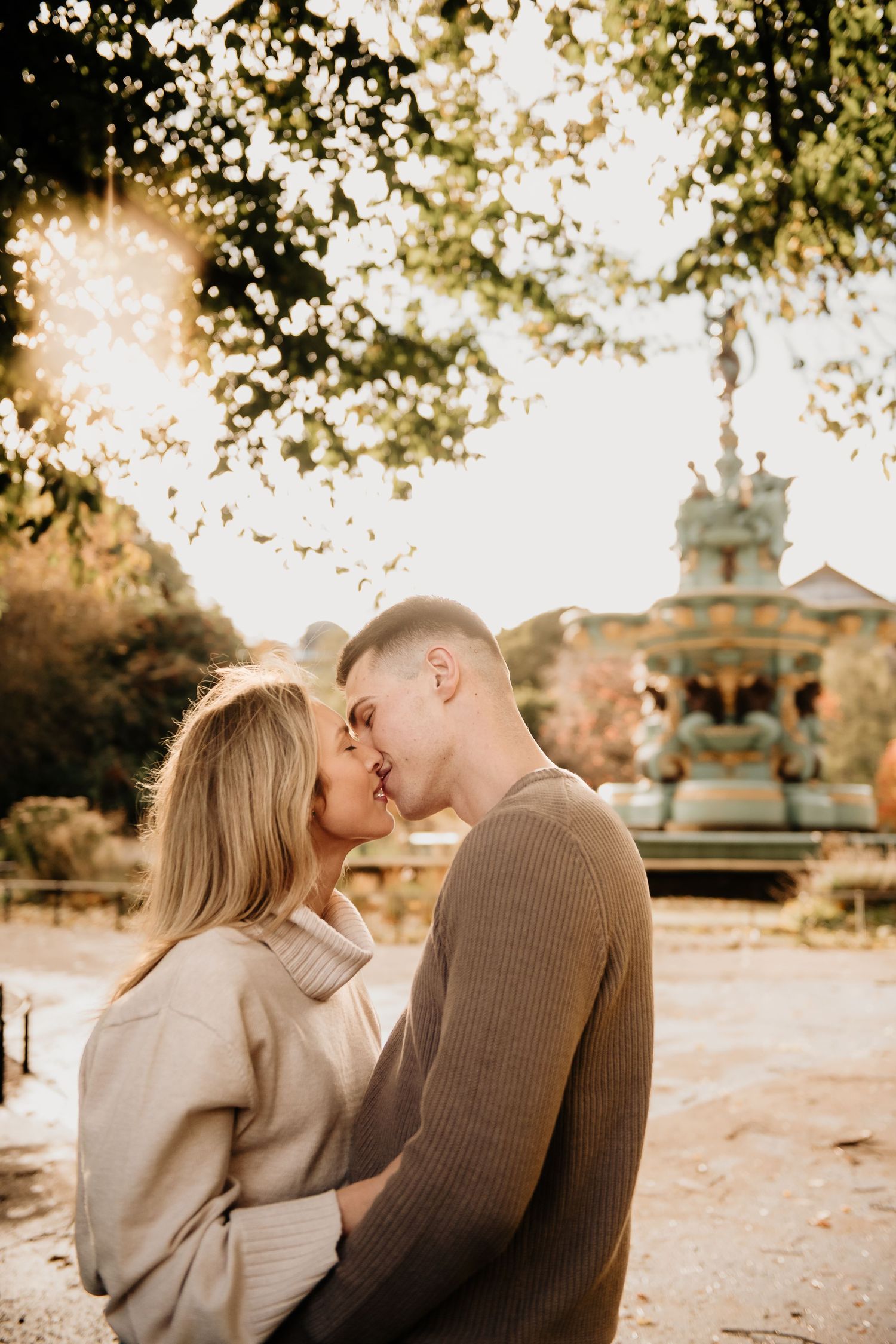 A romantic couple shares tender moments in a park during golden hour with trees and buildings in the Edinburgh background.
