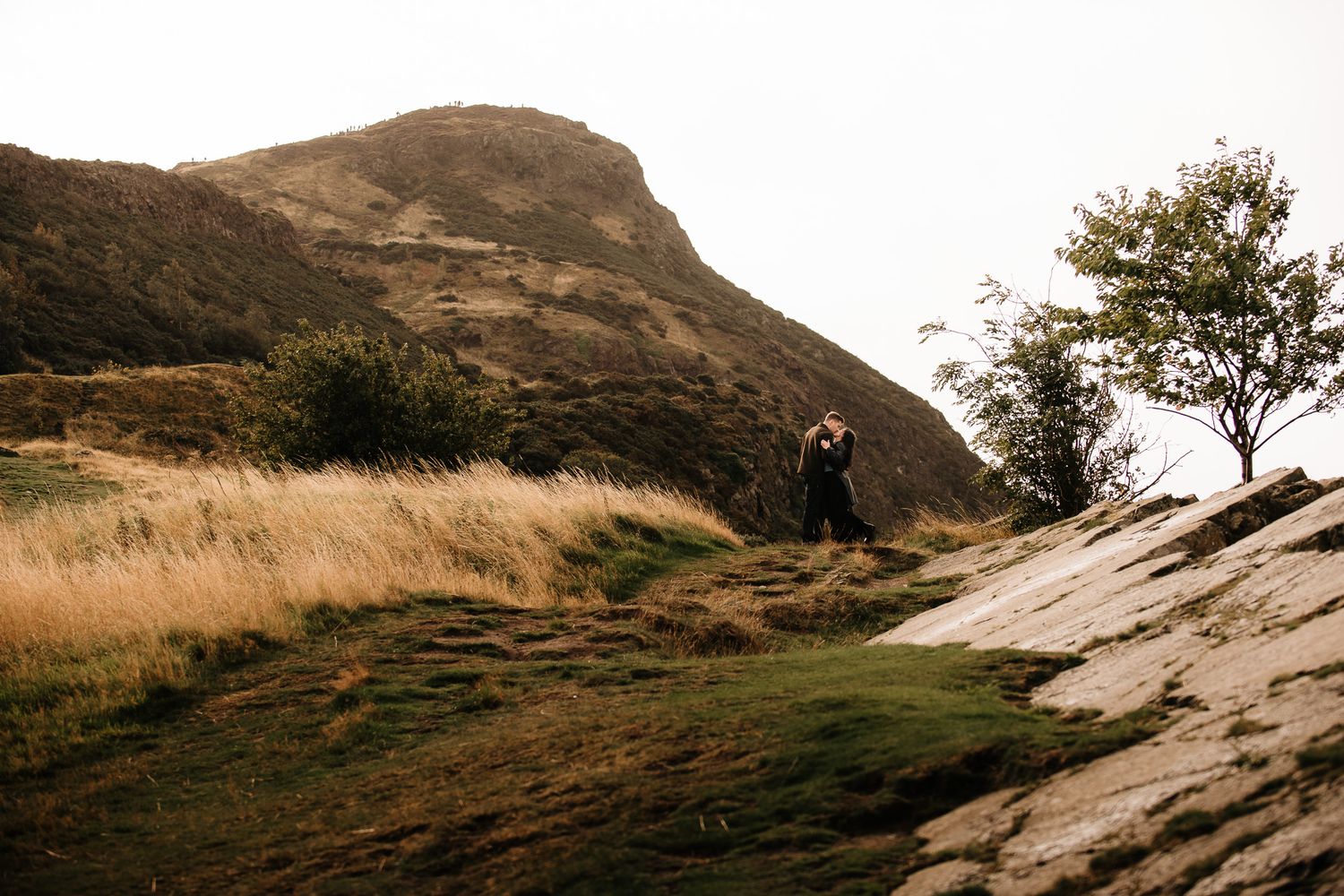 Couple embracing on rocky hillside with golden grass and a lone tree against a dramatic mountain backdrop.