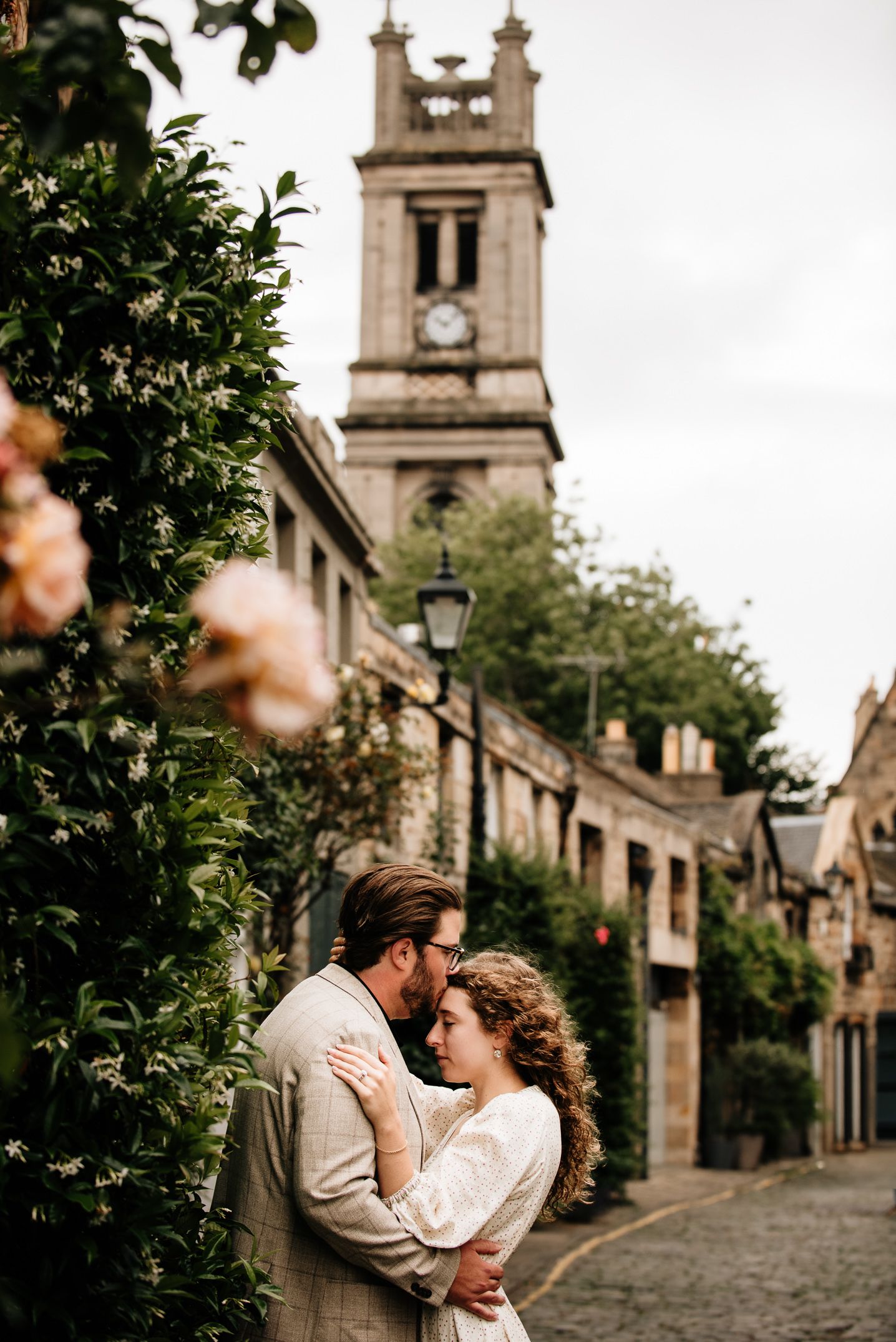 Romantic couple embracing on a cobblestone street beside a historic church tower and blooming roses.