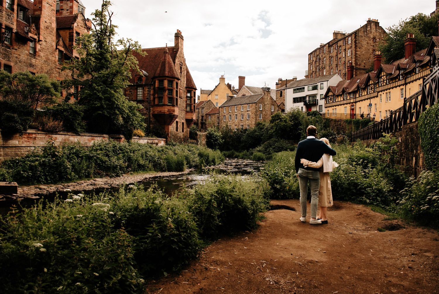 Couple embracing on a path overlooking a lush green valley with historic stone buildings in Edinburgh.