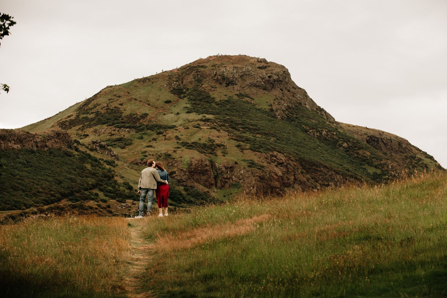 Couple embracing on a grassy trail with a scenic green mountain peak in the background under an overcast sky.