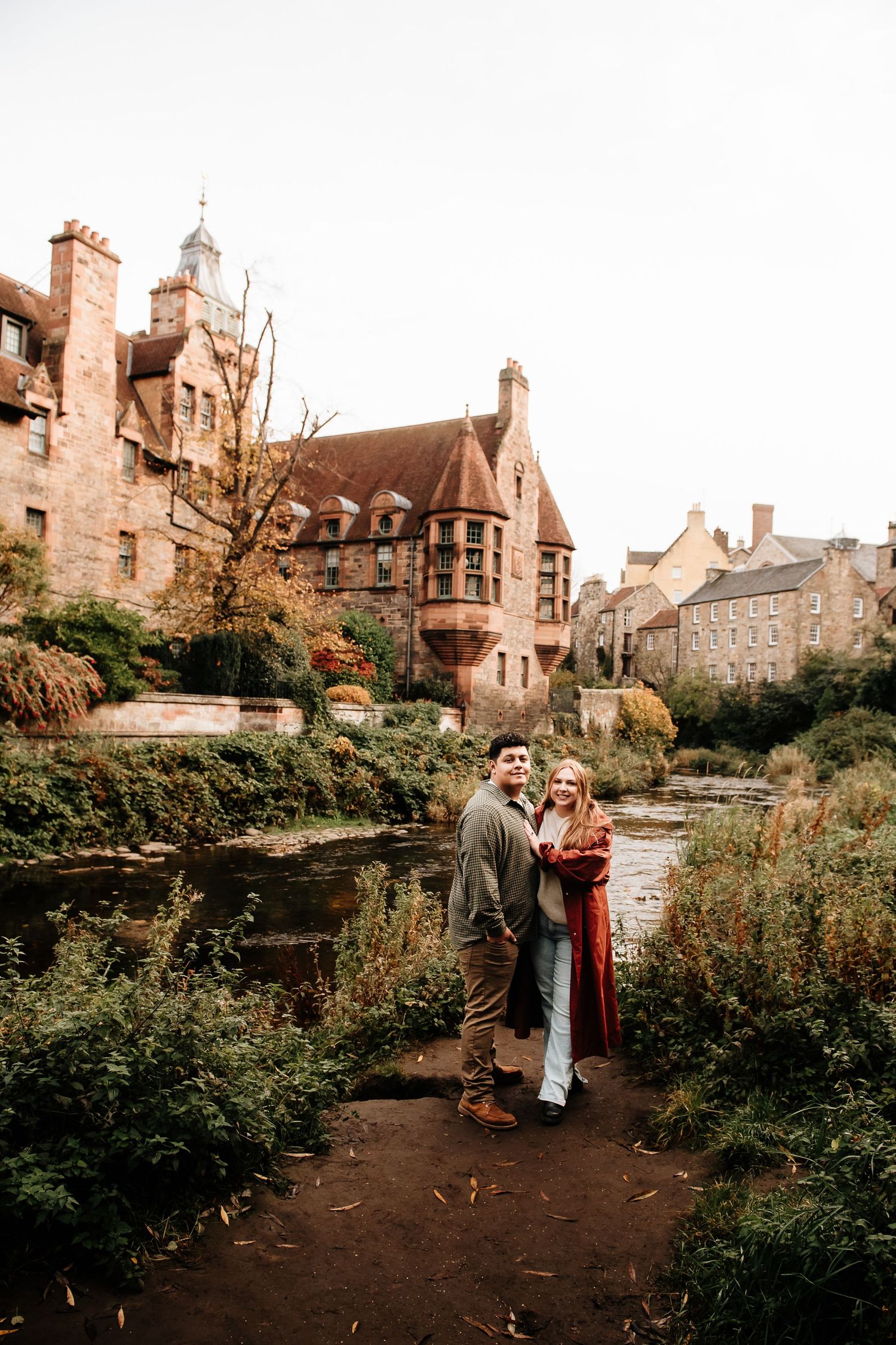 Romantic couple embraces in a garden path with historic stone buildings and autumn foliage in background.