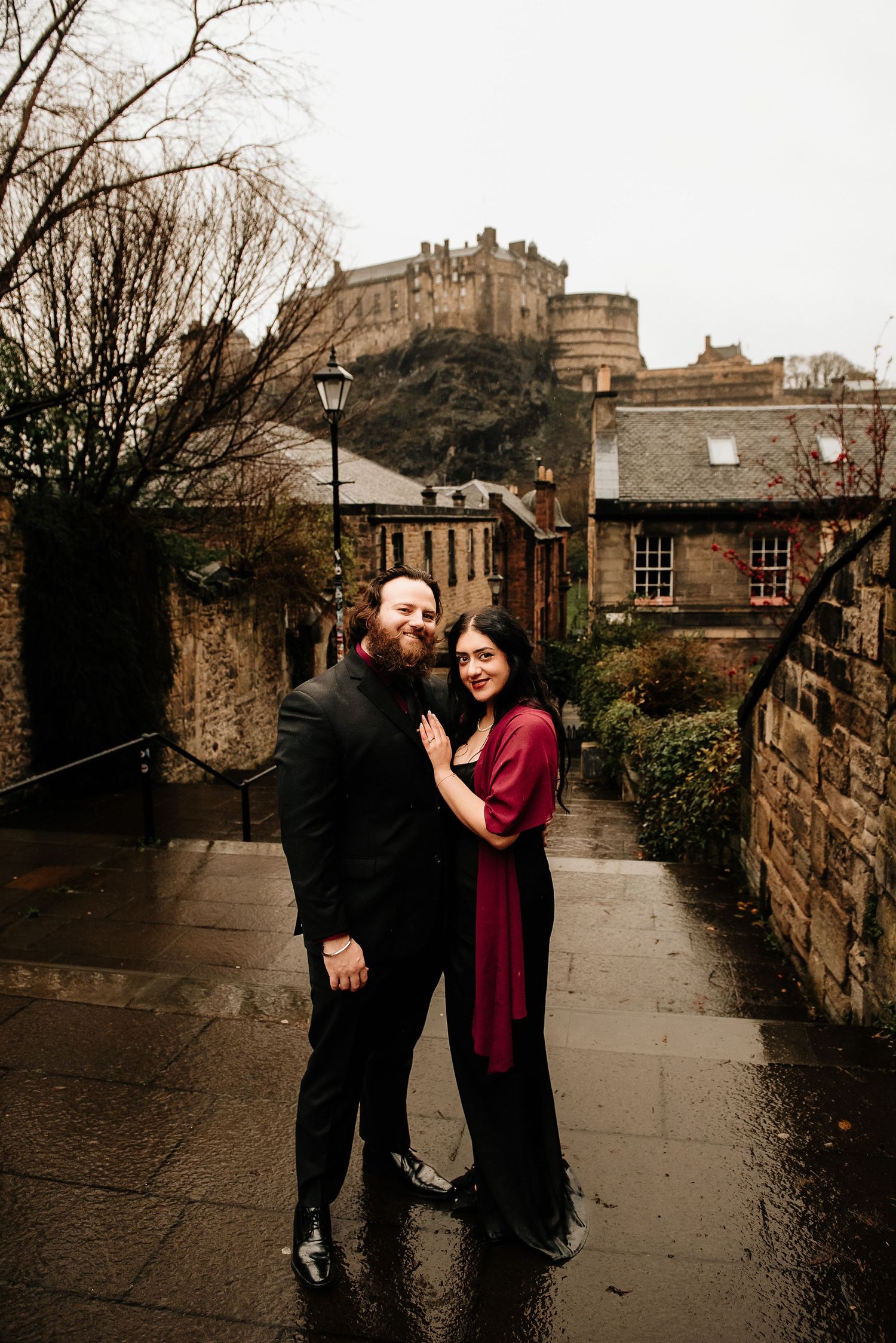 A couple poses for a romantic portrait on wet cobblestone steps with Edinburgh Castle in view.