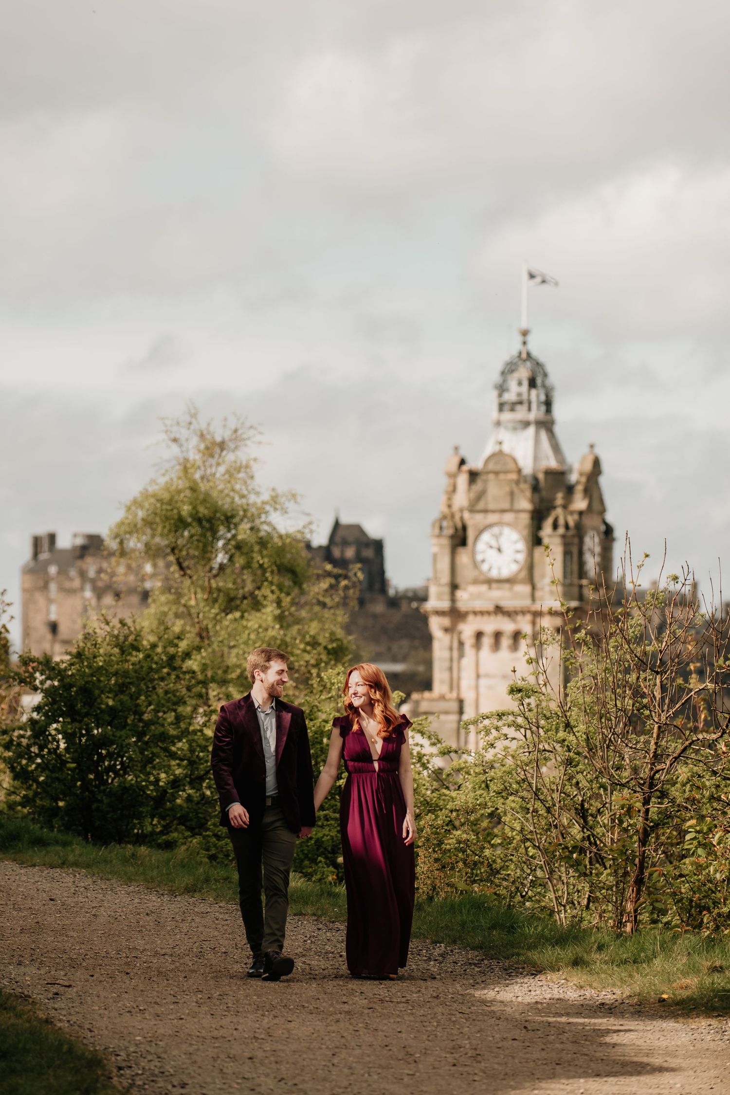 A couple walks along a hilltop path with Calton Hill with Edinburgh's iconic Balmoral Hotel clock tower in the background.