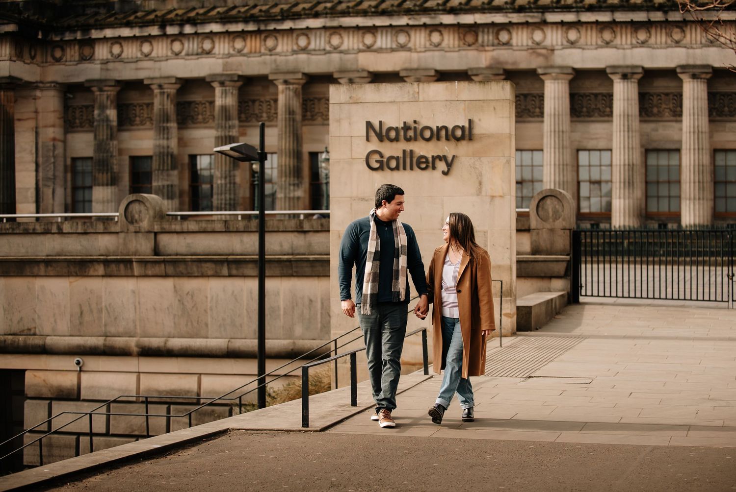 A couple walks along a historic stone building exterior in Edinburgh with ornate architectural details in sepia tones.