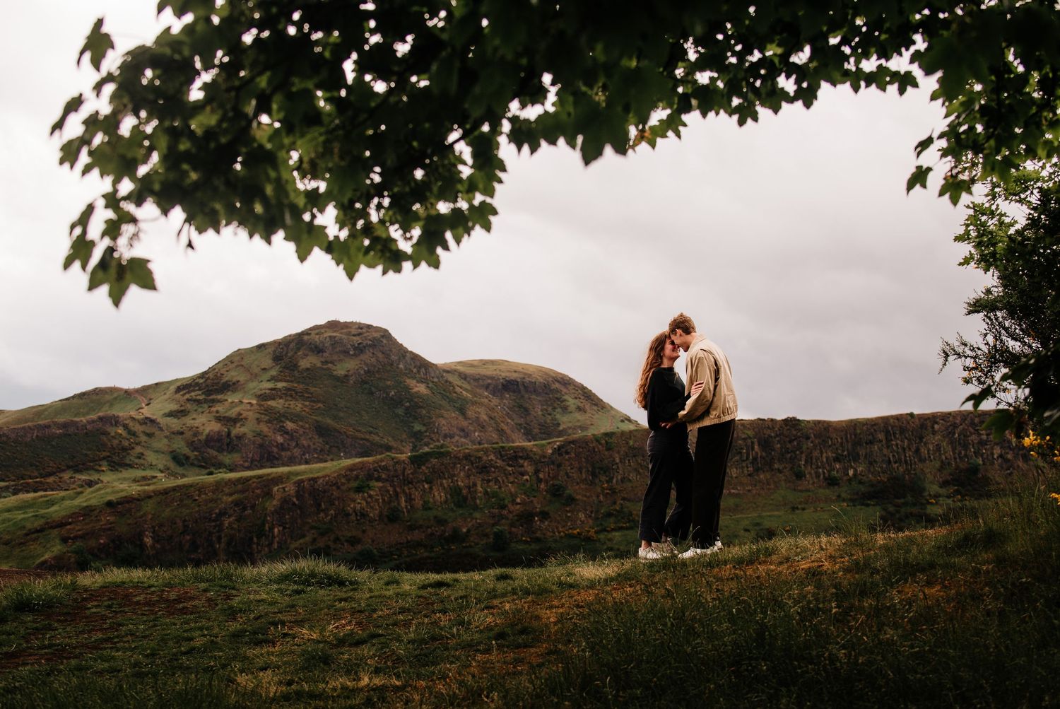 Couple embracing romantically under a tree with Arthur's Seat hill in Edinburgh, Scotland backdrop on a cloudy day.