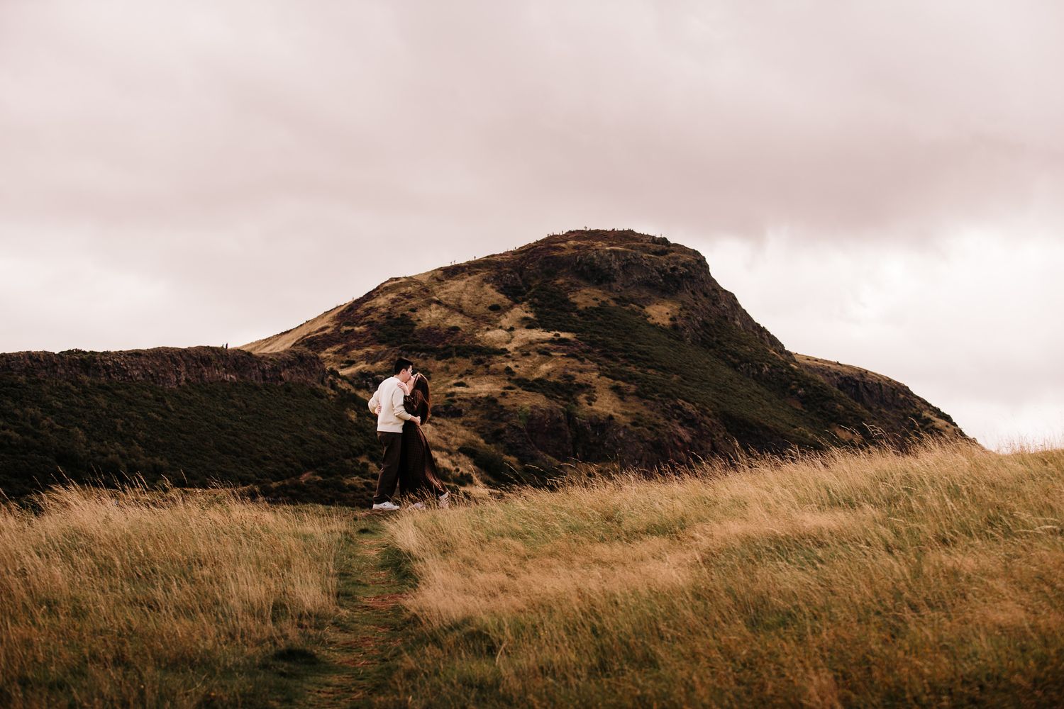 Couple embracing on a scenic highland trail with dramatic hills and golden grass under a cloudy sky.