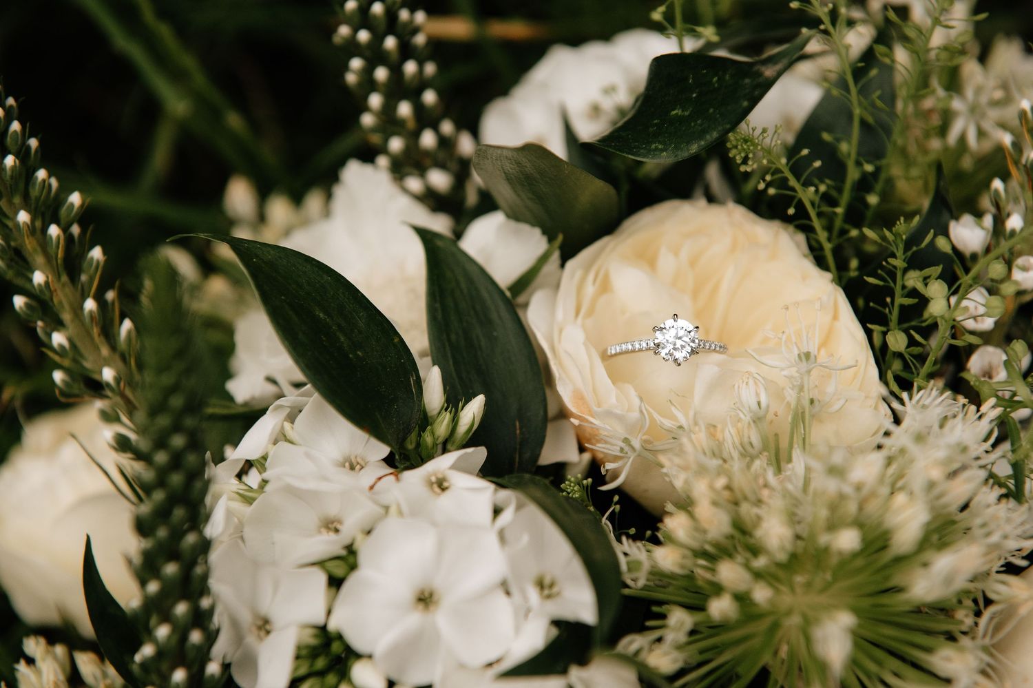 Close-up of a delicate wedding bouquet with white flowers, greenery and a diamond engagement ring.