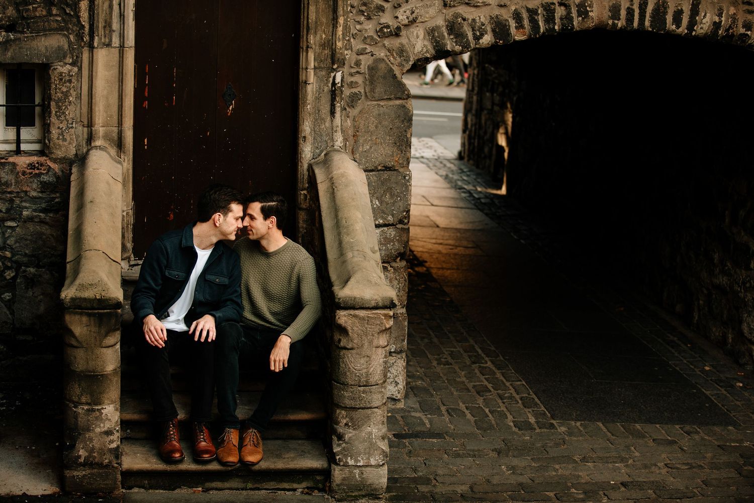 Two people sit on stone steps in an atmospheric alleyway with dark archways and traditional architecture.