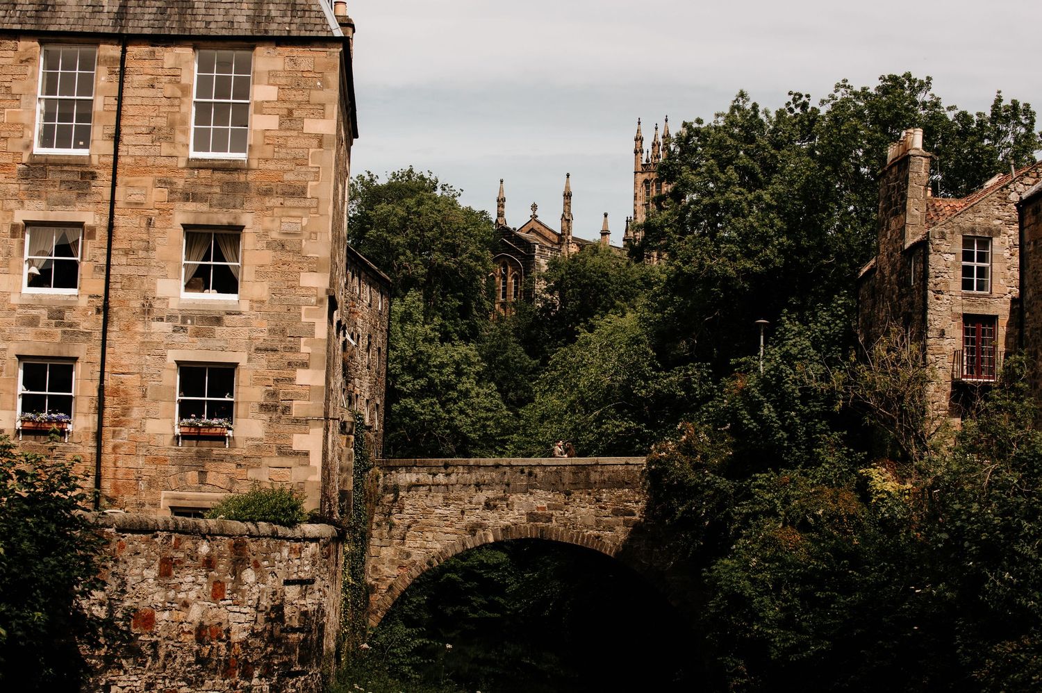 Historic stone bridge and building in Edinburgh's Dean Village, surrounded by lush green trees with Gothic spires visible.
