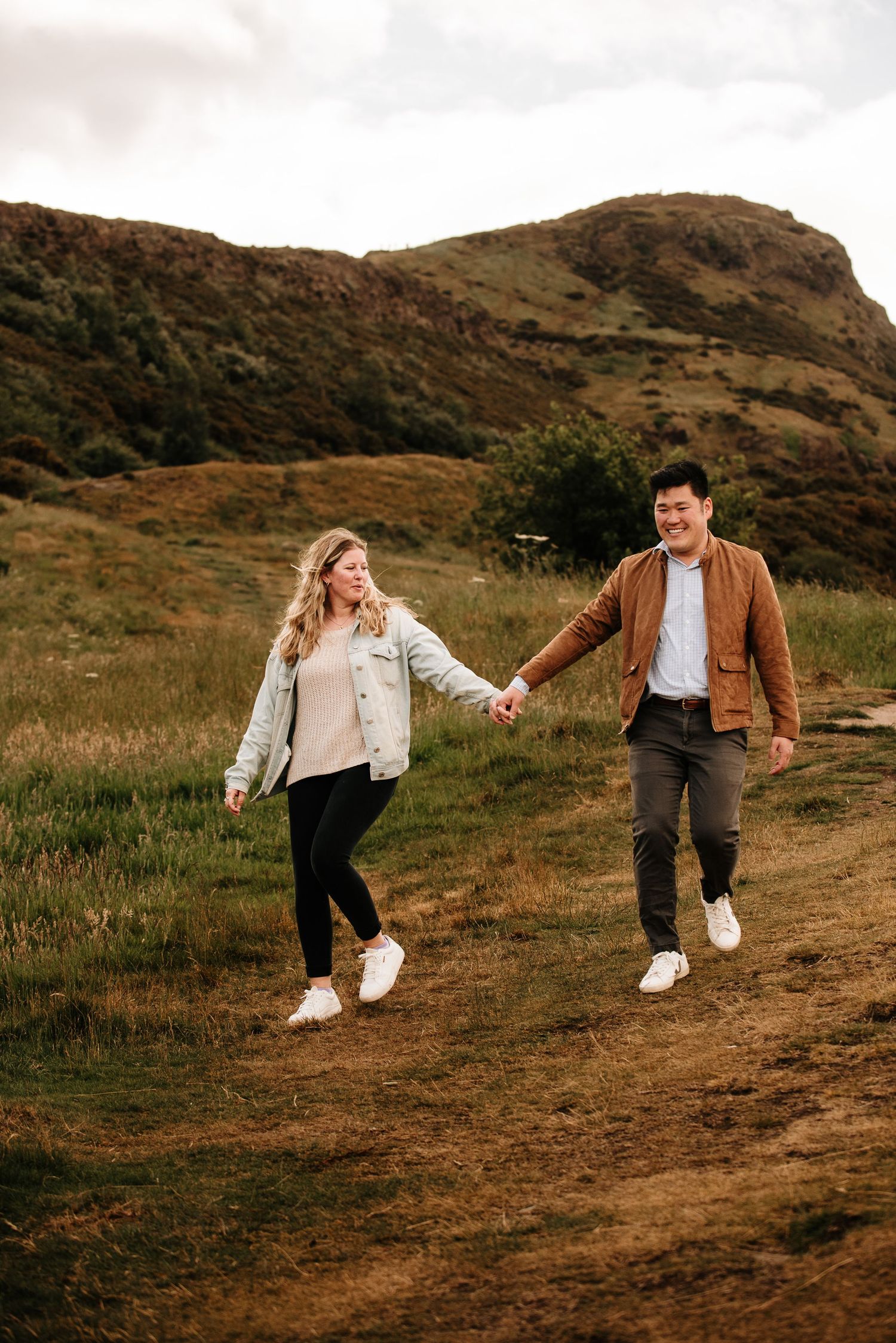 A couple holds hands while walking along a dirt trail on a hillside.