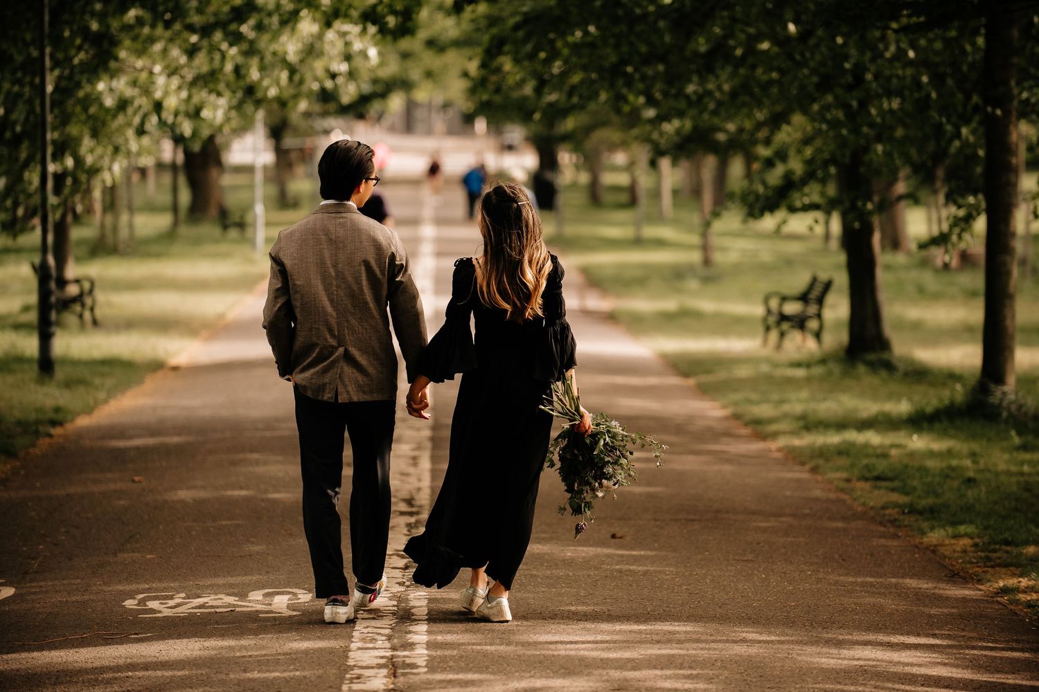 A couple walks hand in hand down a tree-lined path in a park at sunset, with the person on the right holding flowers.