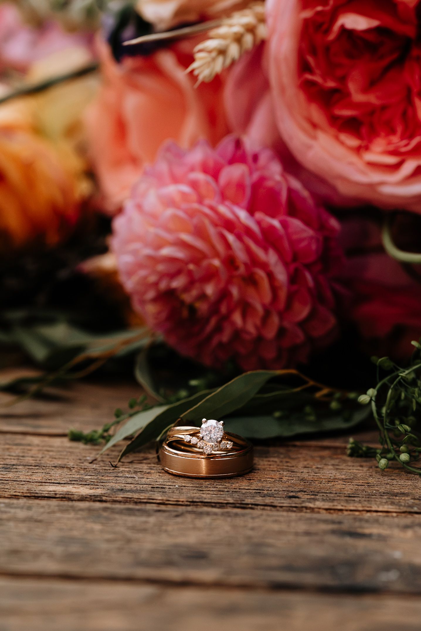 Gold wedding rings rest on rustic wood beside a vibrant bouquet of pink dahlias, coral roses, and greenery.