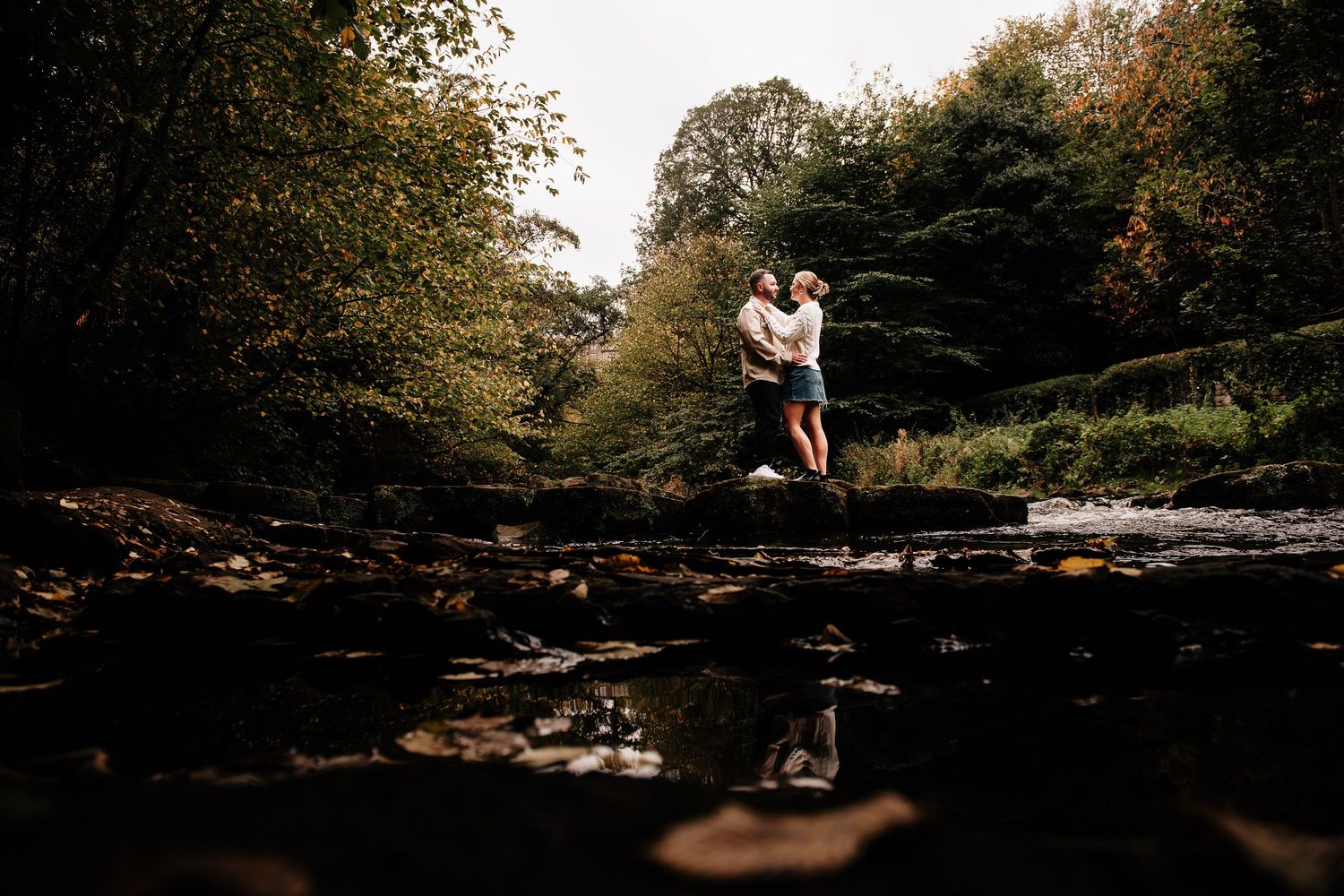 Silhouetted figures standing on rocks by a stream in a wooded setting at sunset.