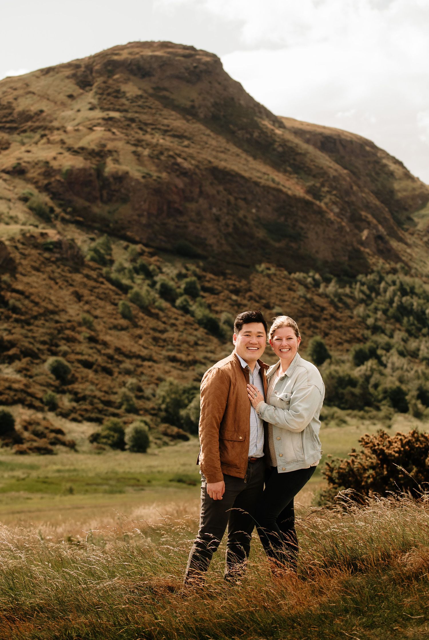 A couple poses together in front of a dramatic brown mountain landscape on a cloudy day.