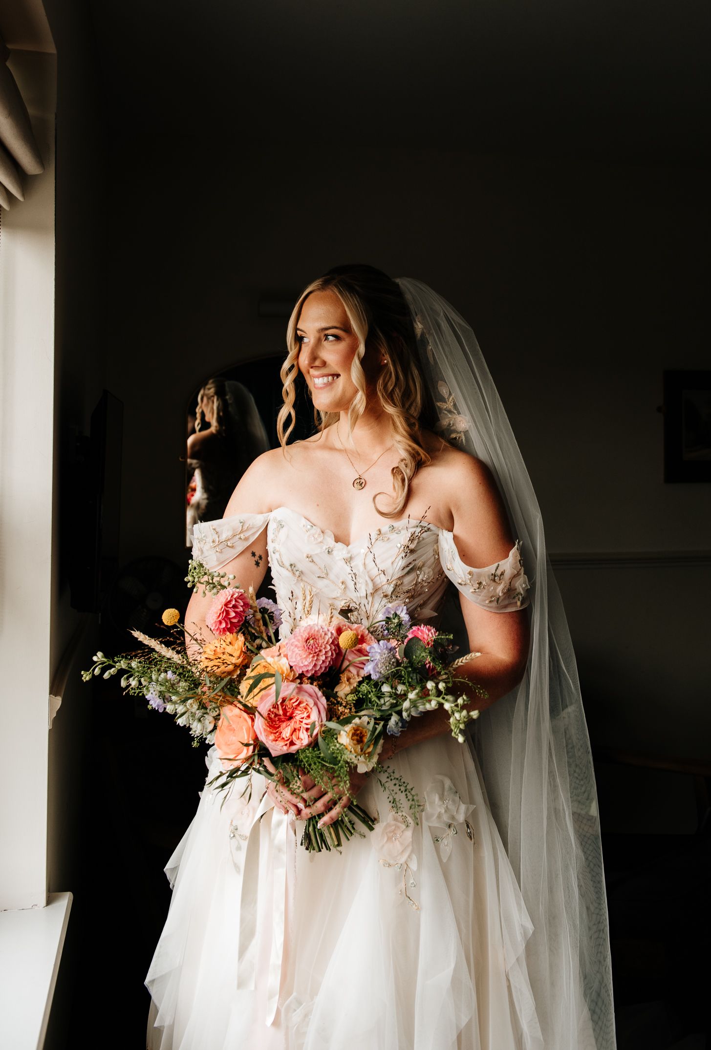 Smiling bride in off-shoulder lace wedding gown holds colorful bouquet with veil, standing by window in soft natural light.