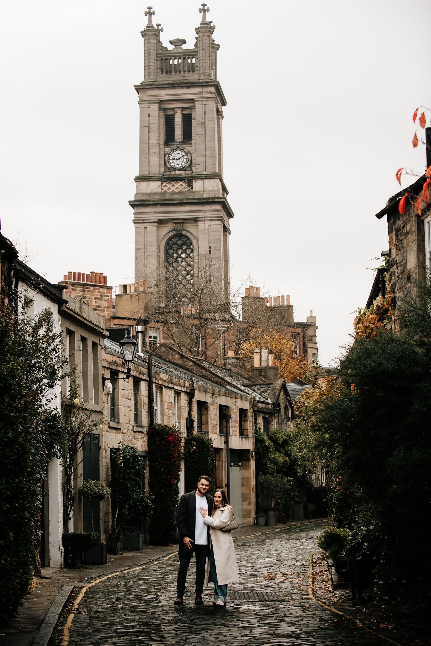 A couple walks along a historic cobblestone street with church spires and stone buildings.
