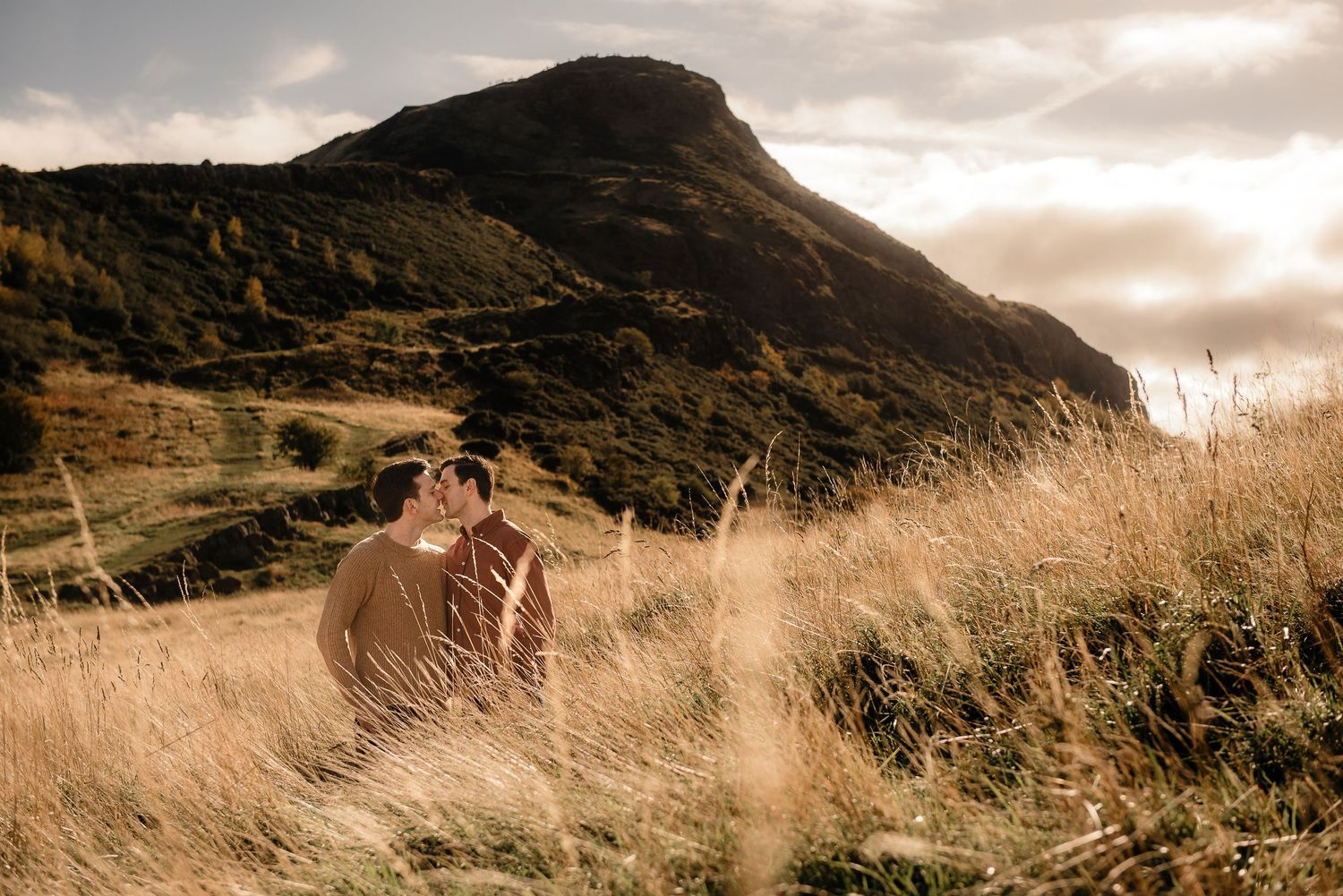 Two people embrace in tall grass with a dramatic mountain landscape at sunset.