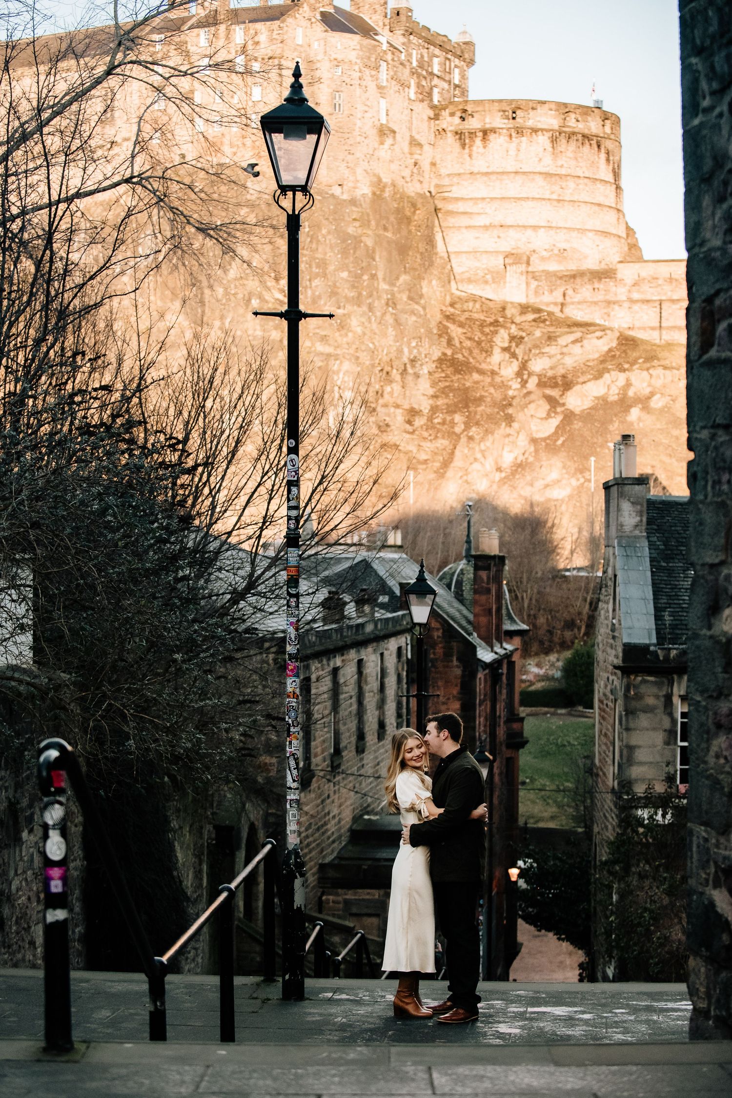 Atmospheric evening scene along a lamp-lit path beneath Edinburgh Castle's dramatic cliffs.