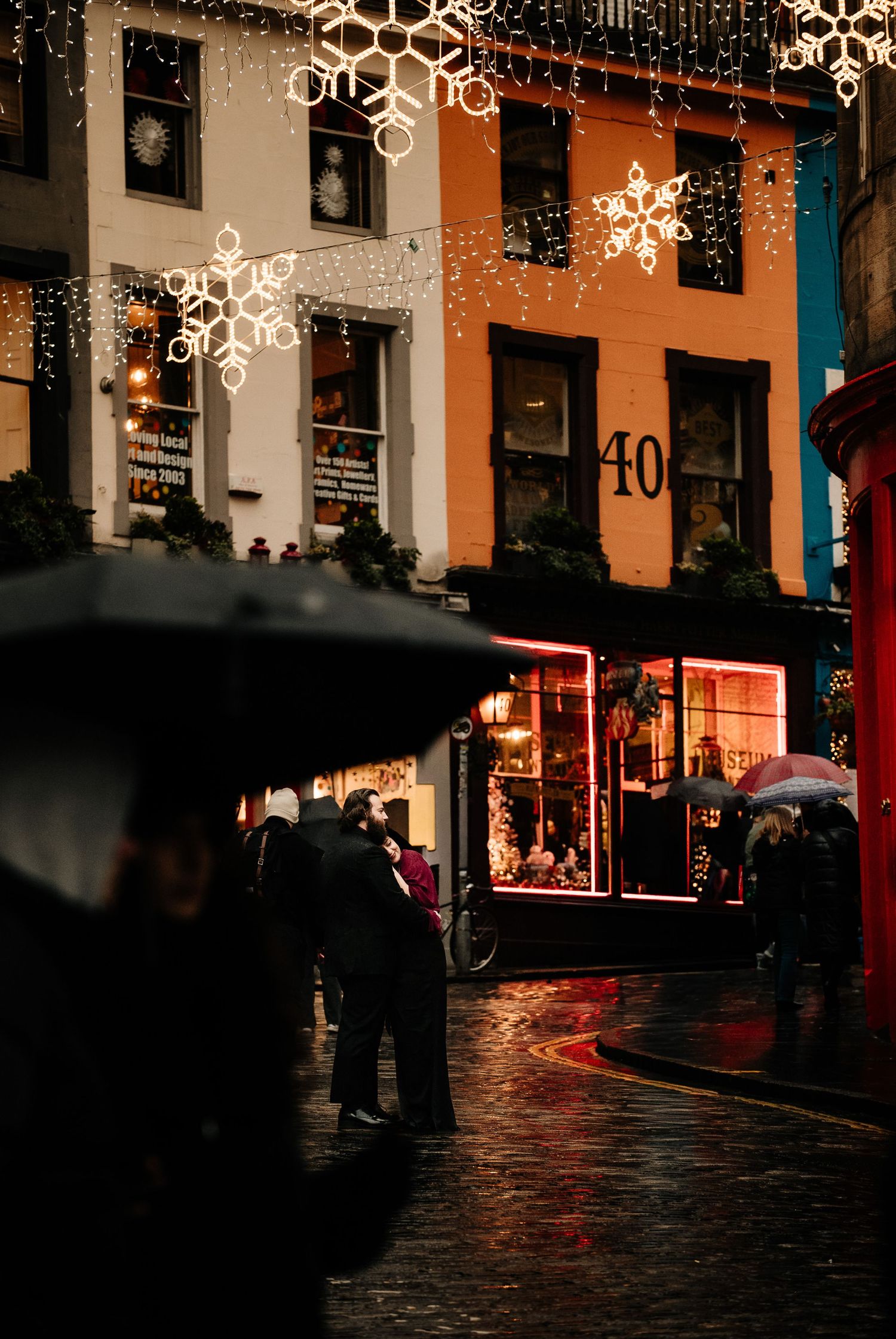 Illuminated winter street scene with hanging snowflake decorations and glowing storefronts reflecting on wet cobblestones.