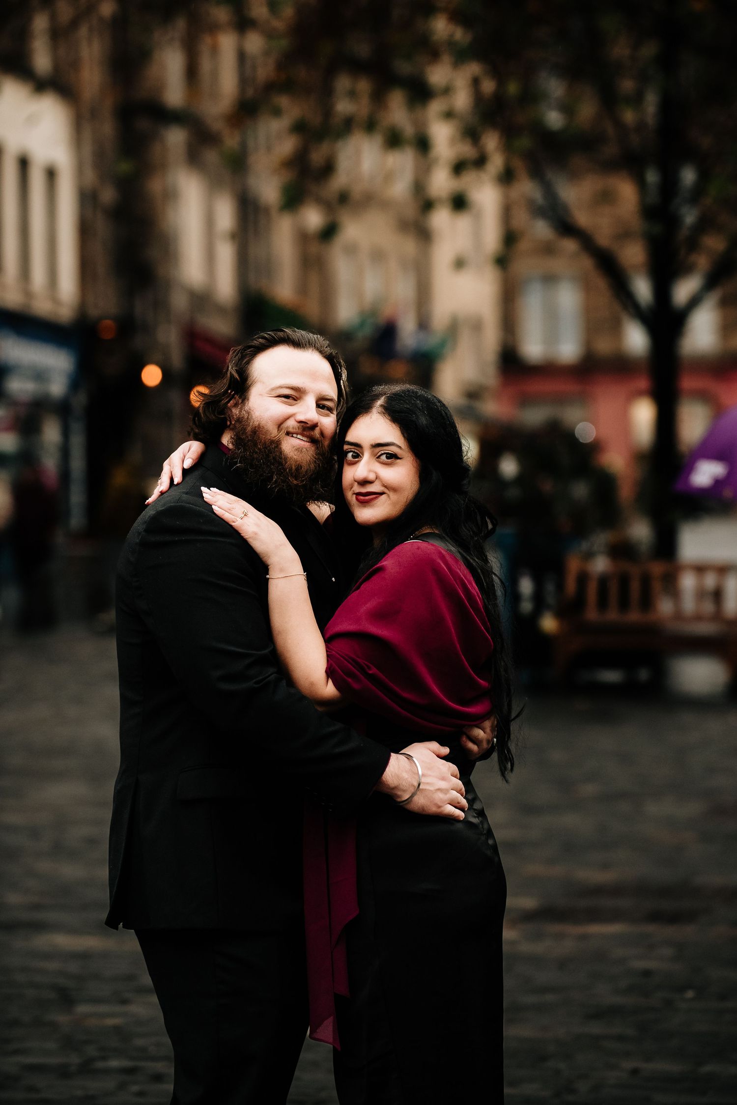 A couple embraces on a romantic cobblestone street at dusk, with the woman wearing a burgundy wrap and black dress.