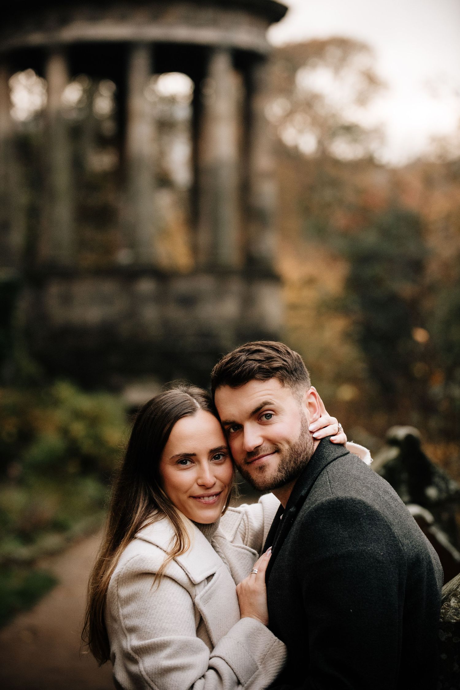 A couple shares a tender moment near a historic stone gazebo in an autumn garden.