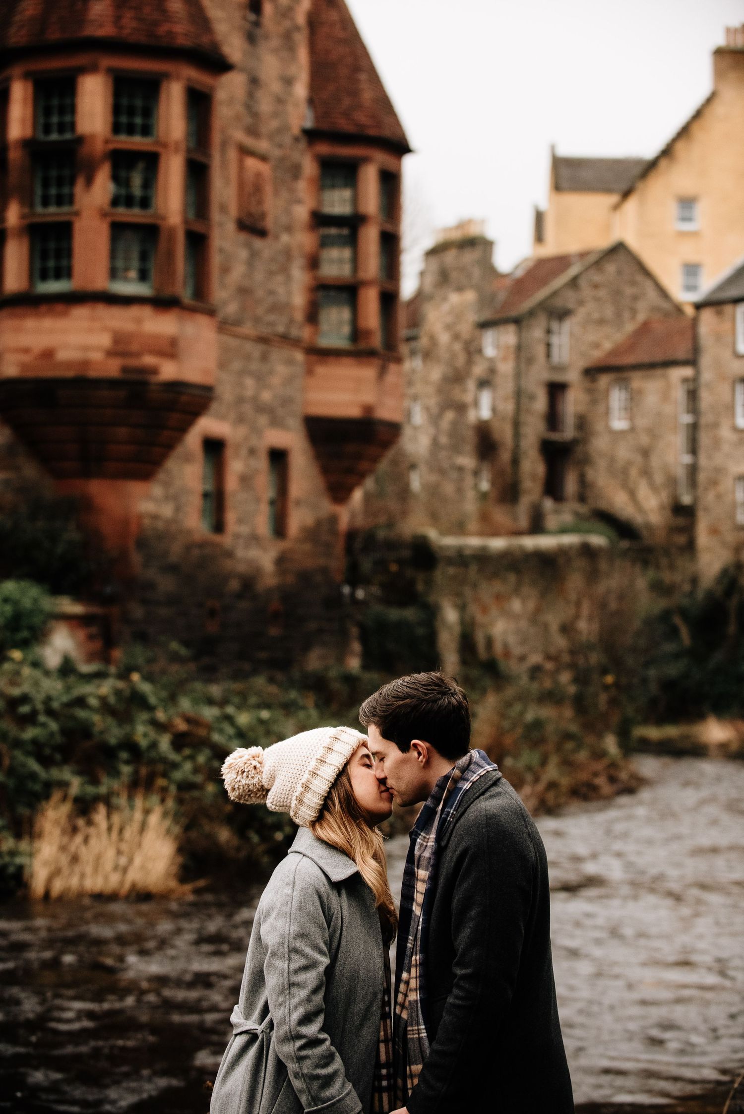 A couple embraces near historic stone buildings on a winter day in Edinburgh.