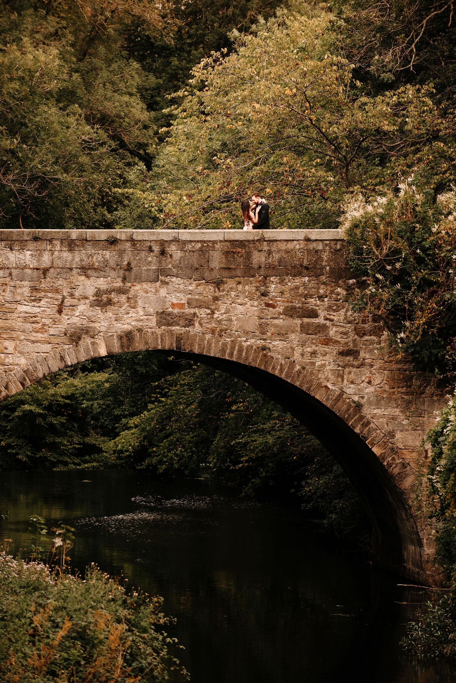 A stone bridge arch frames a romantic moment in a lush natural setting.