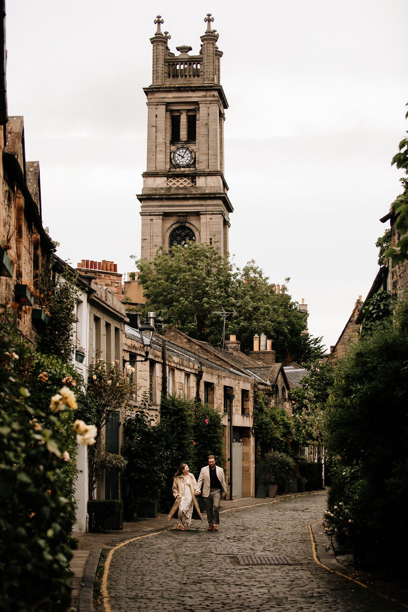 Couple walking hand in hand on a cobblestone street with a historic clock tower in Edinburgh, Scotland.