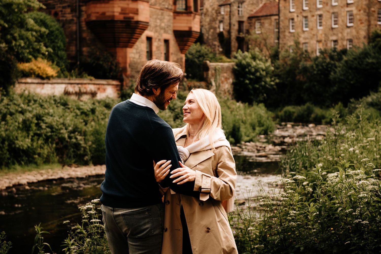 Couple embracing and smiling at each other beside a scenic river with historic stone buildings in background.