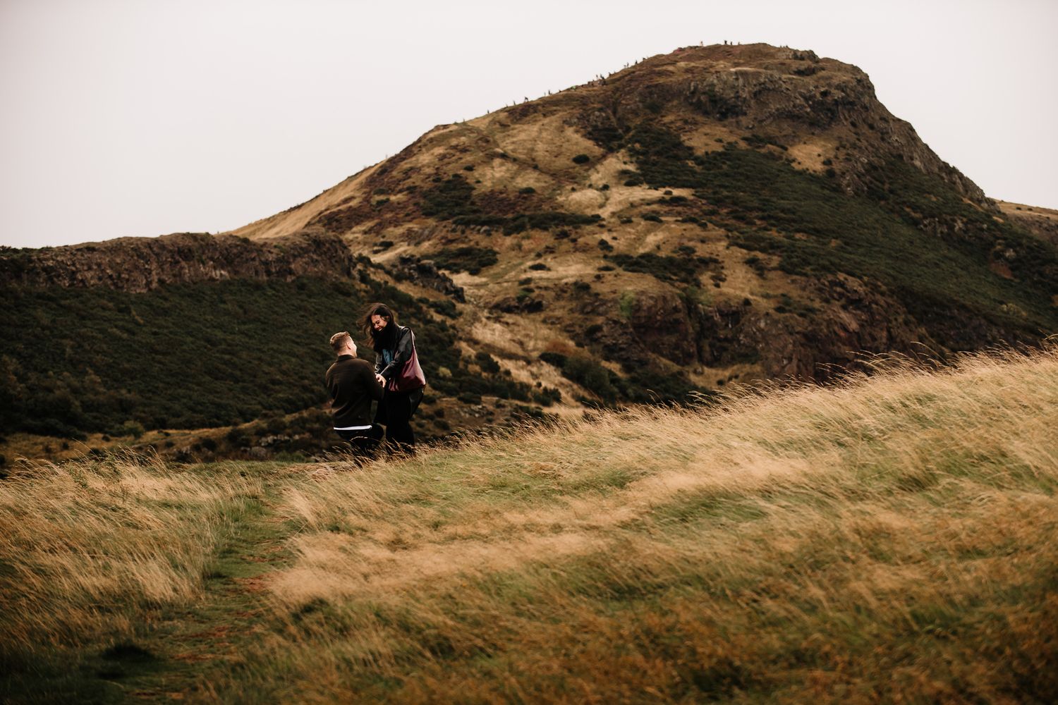 Two hikers walking through golden grasslands with a rugged hill in the background on an overcast day.