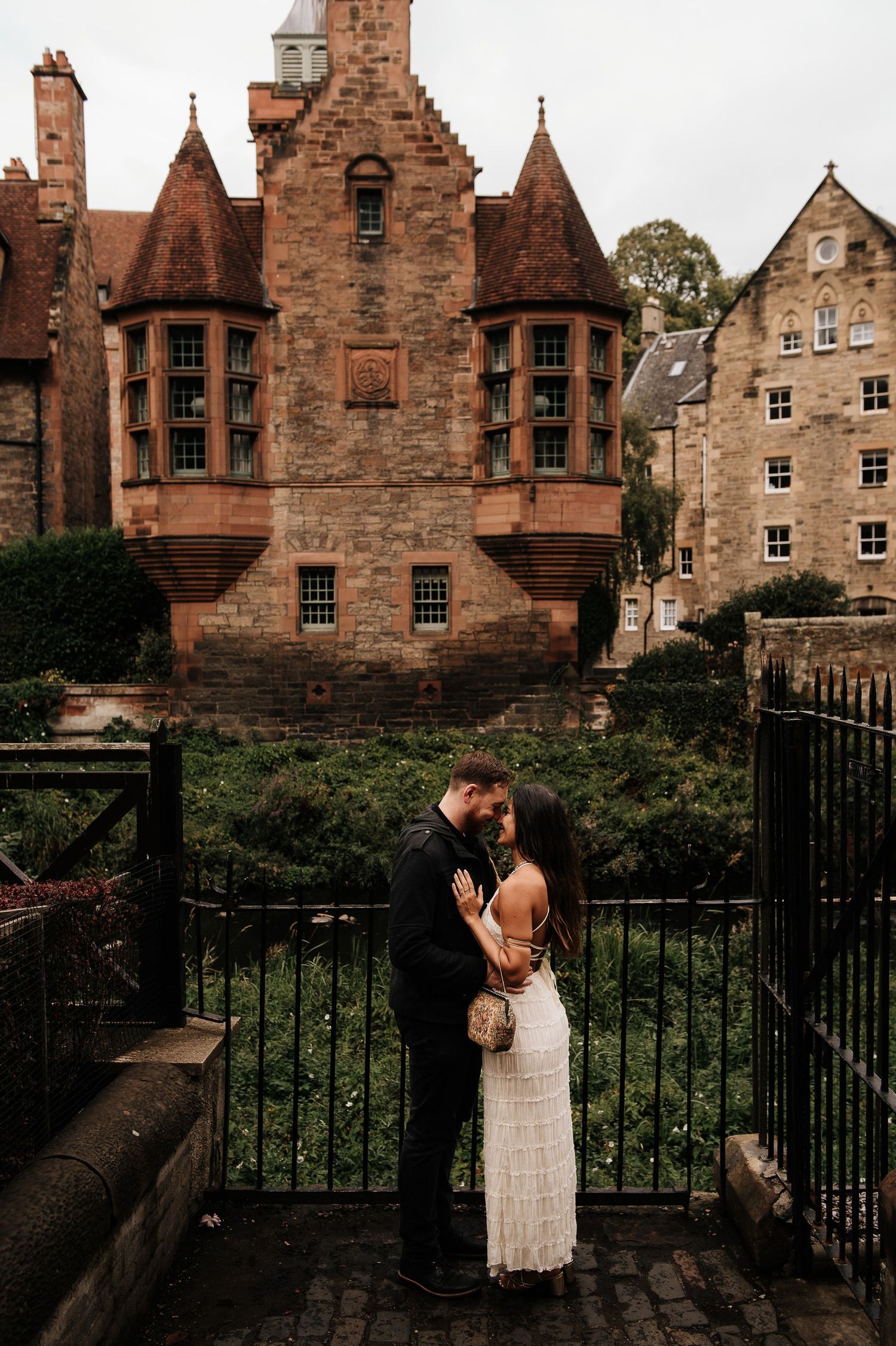 Couple shares romantic moment along iron railing with historic stone buildings behind them.
