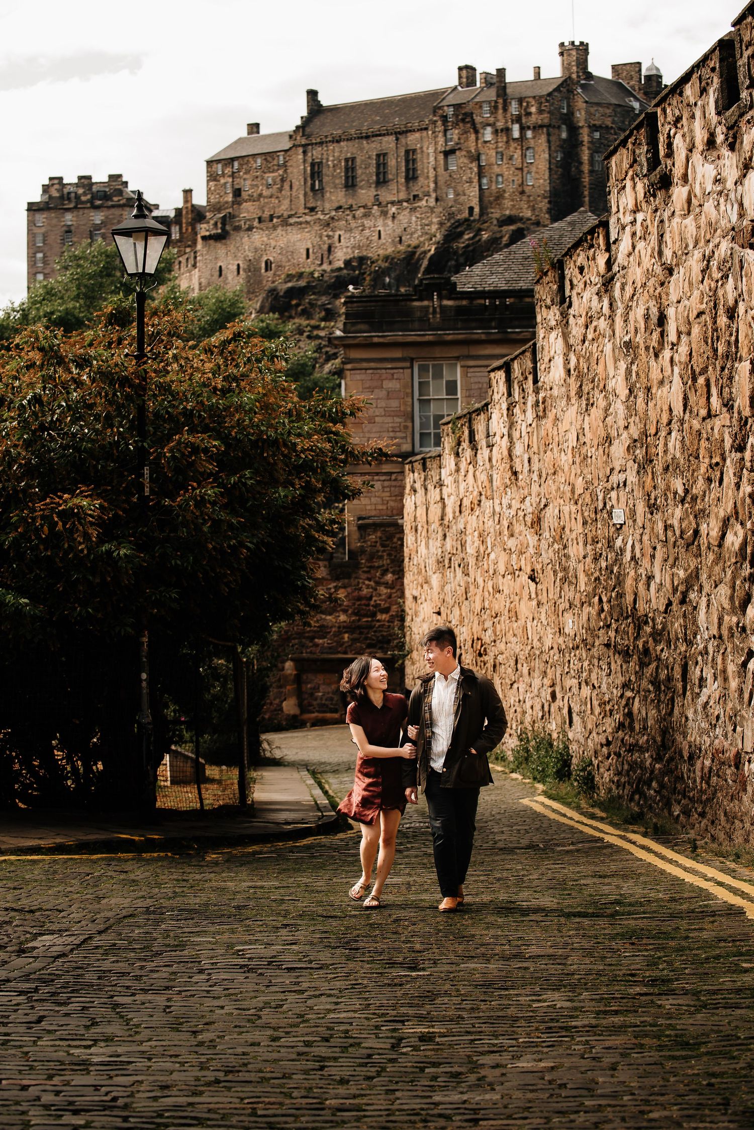 Couple walking hand-in-hand on cobblestone path with Edinburgh Castle in background.