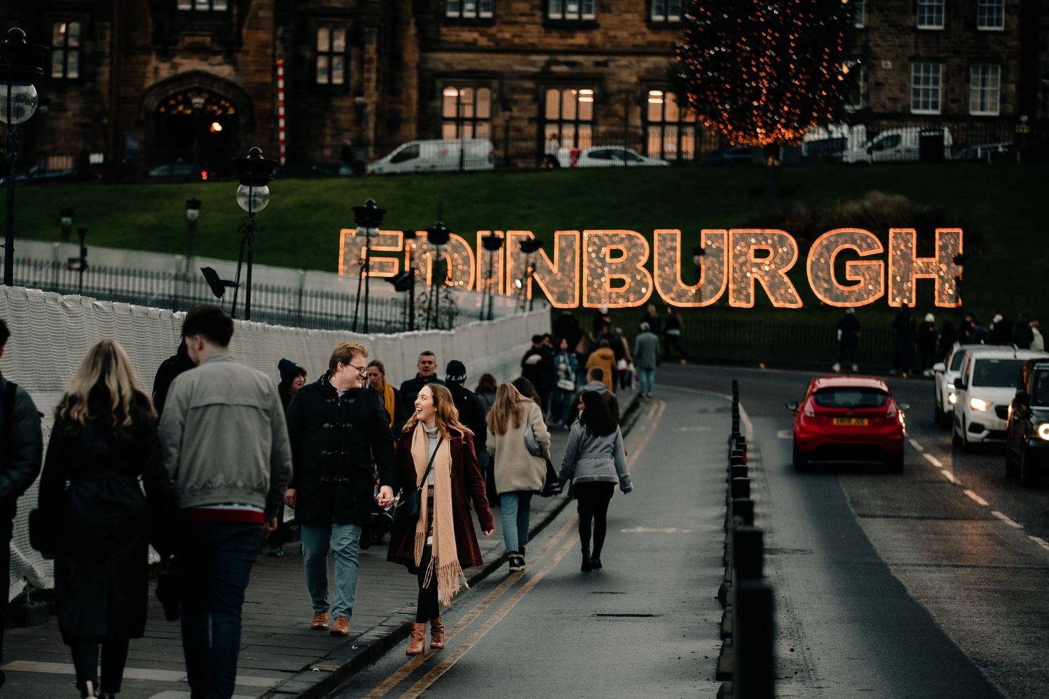 Illuminated Edinburgh sign glows against twilight sky as pedestrians walk along city street.
