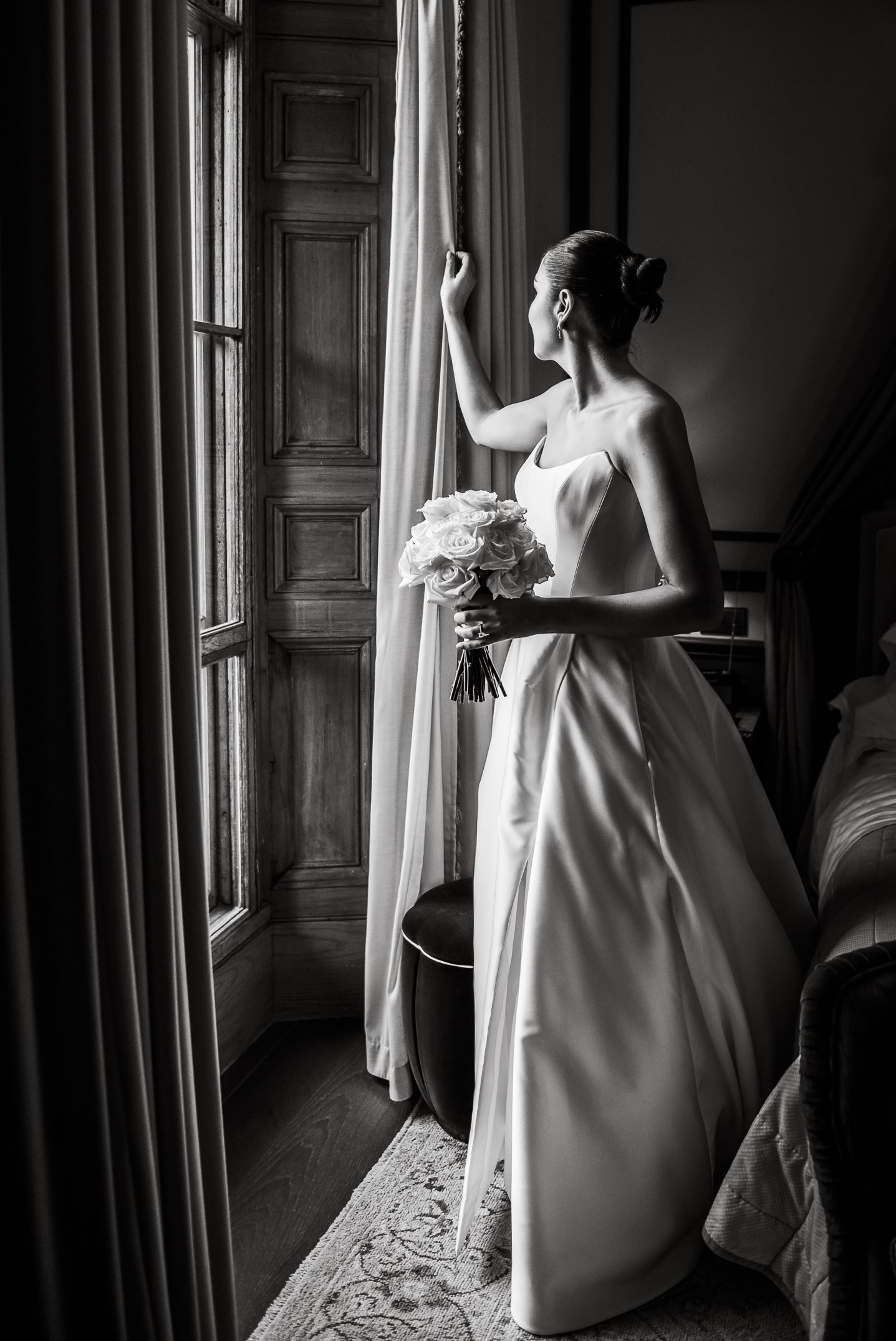Elegant figure in white dress stands by window in dramatic black and white composition.