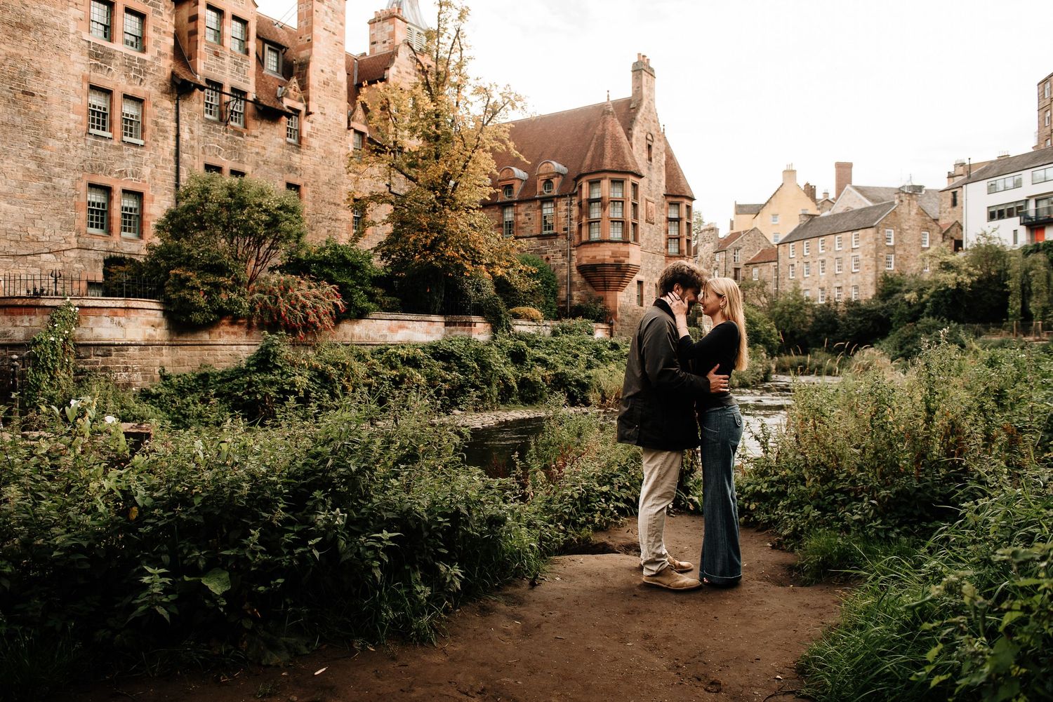 A couple shares a romantic moment in a garden setting surrounded by lush greenery and brick buildings in Dean Village