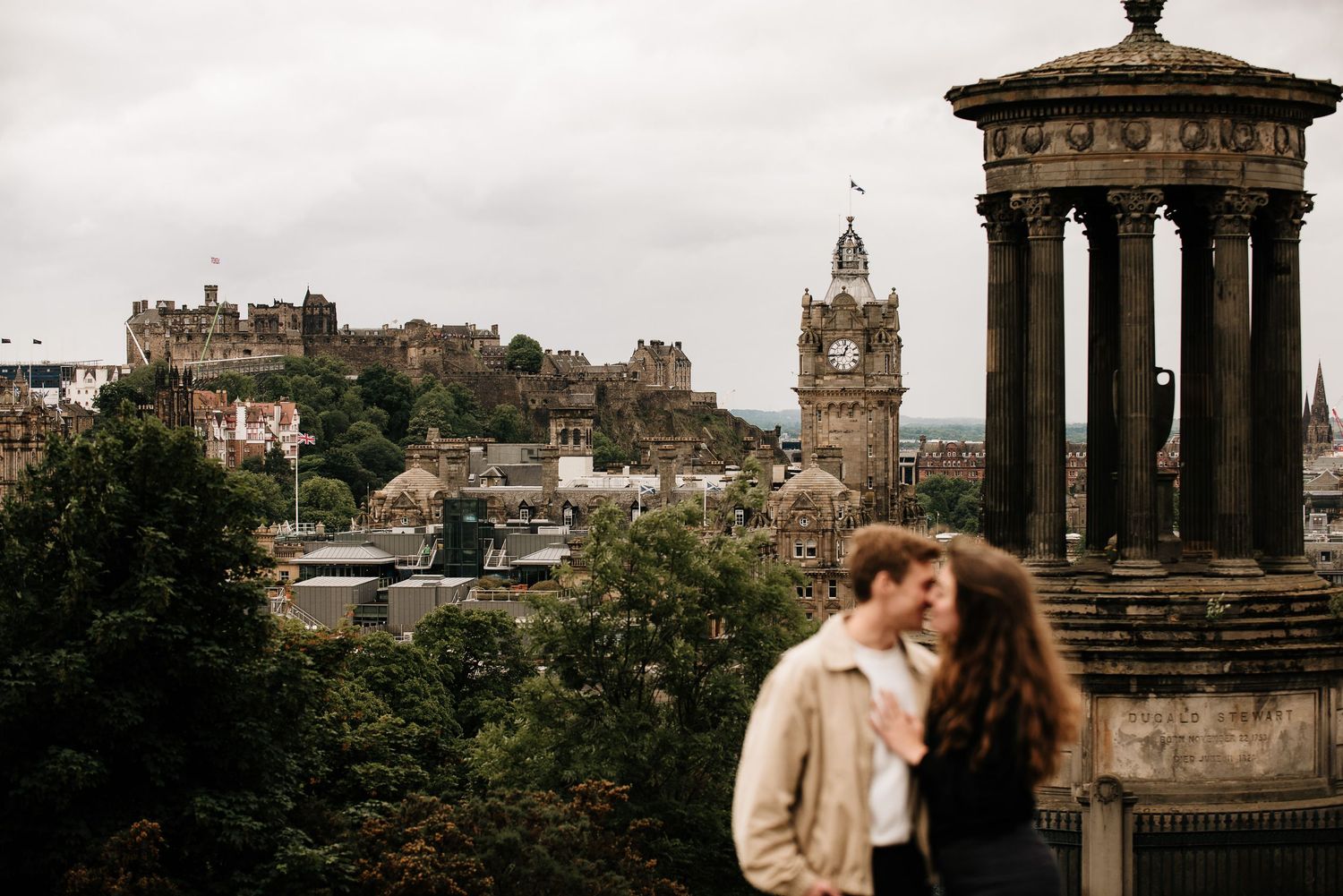 Couple sharing a romantic moment at Calton Hill with Edinburgh Castle and Balmoral Clock Tower in the background.