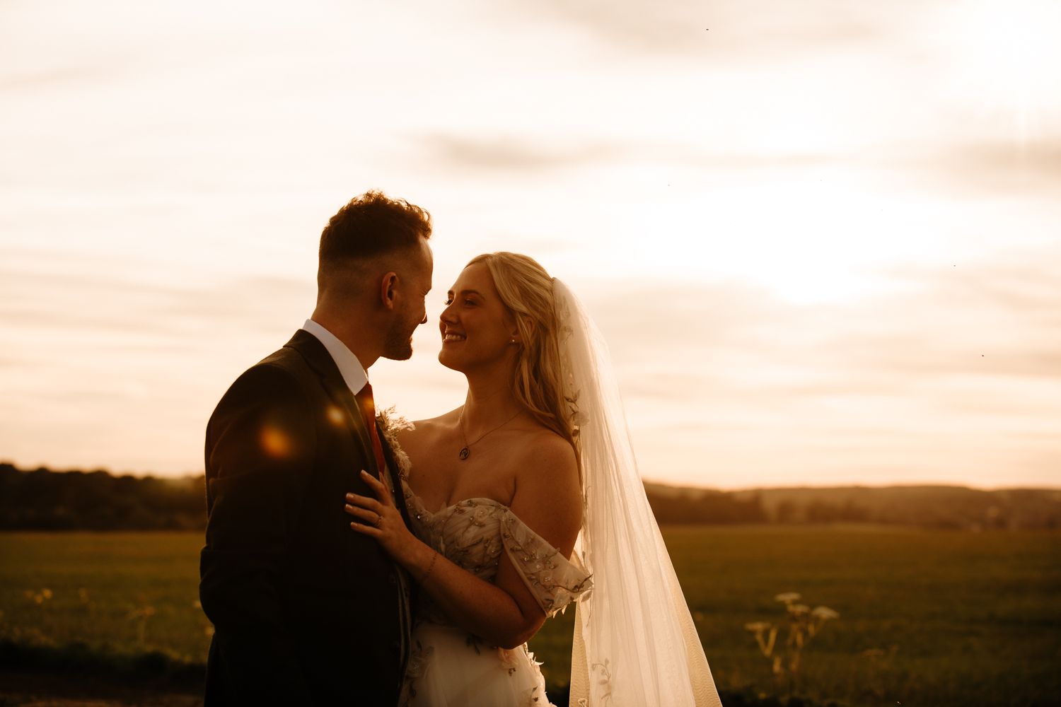 Bride and groom share a romantic moment at golden sunset in an open field, bride wearing lace gown and veil.