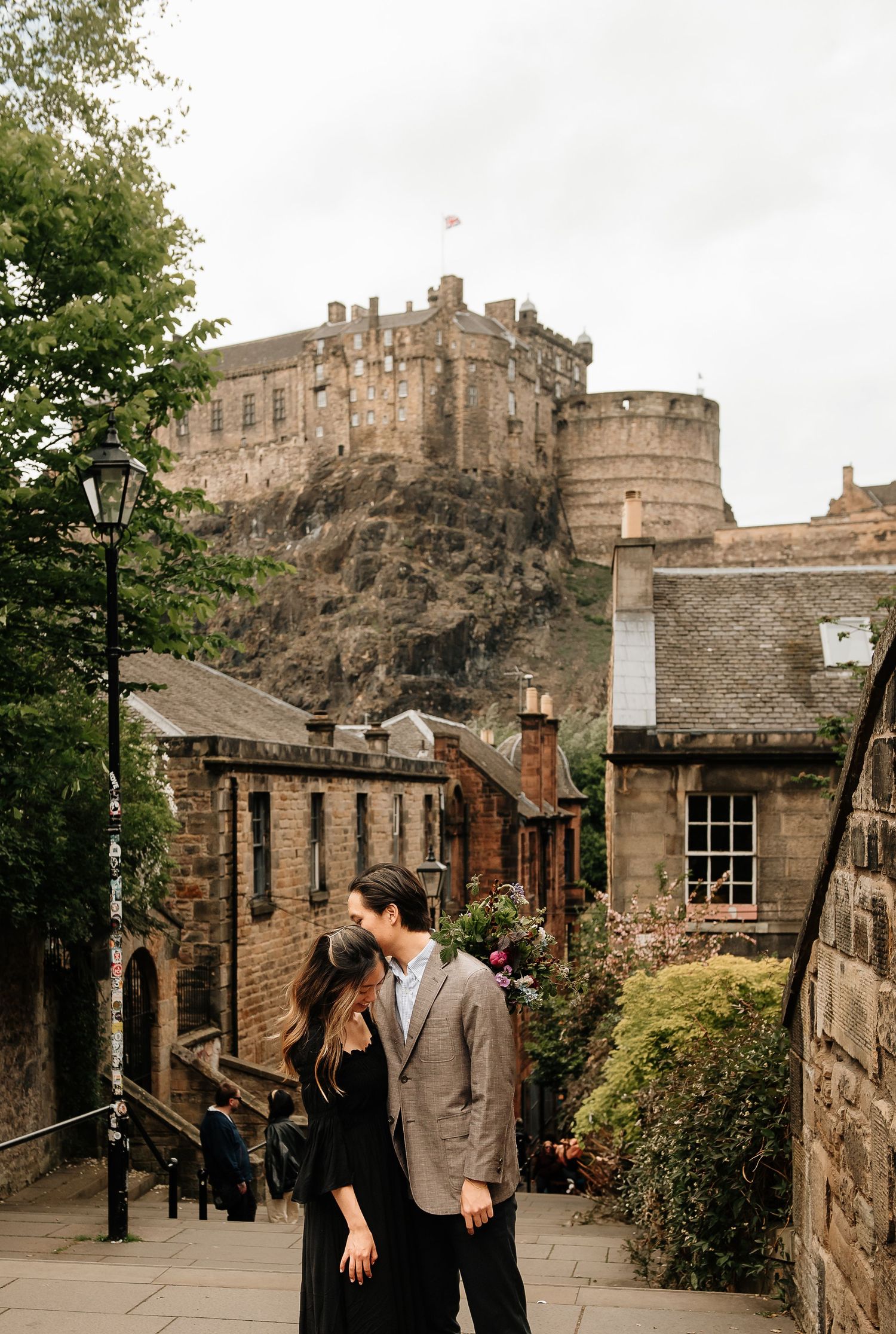 A romantic couple embraces on historic steps with Edinburgh Castle towering above.