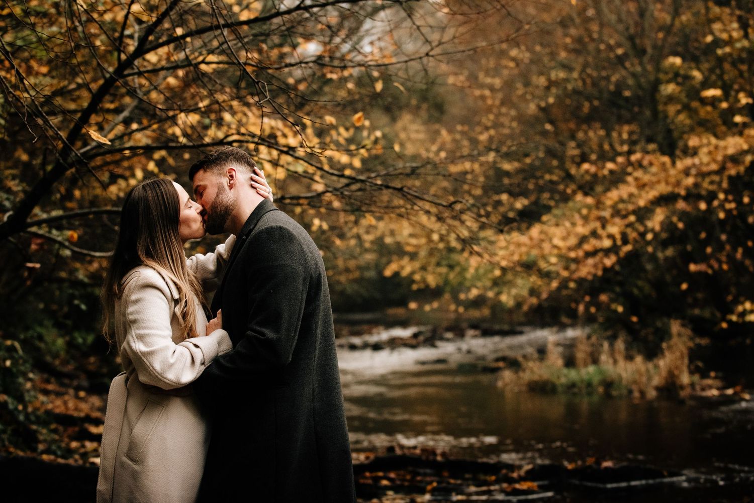 Couple embracing under golden autumn trees near a tranquil stream.