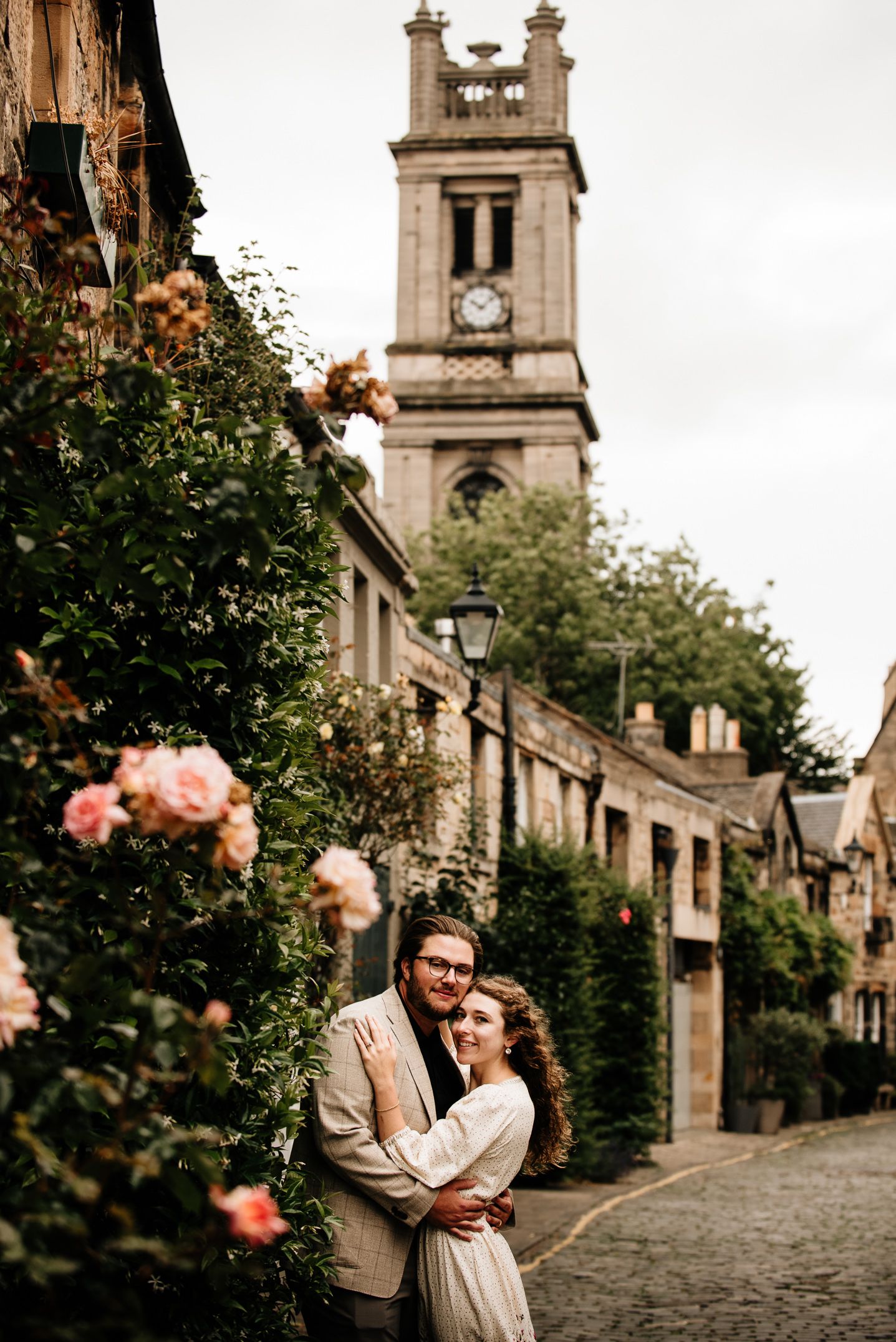 Couple embracing on cobblestone street with blooming roses and historic church tower in Edinburgh, Scotland.