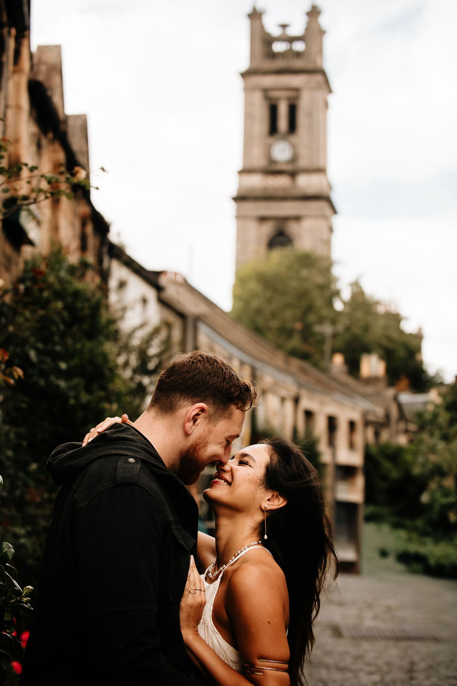 A couple shares an intimate moment on cobblestone streets with a historic church tower backdrop.