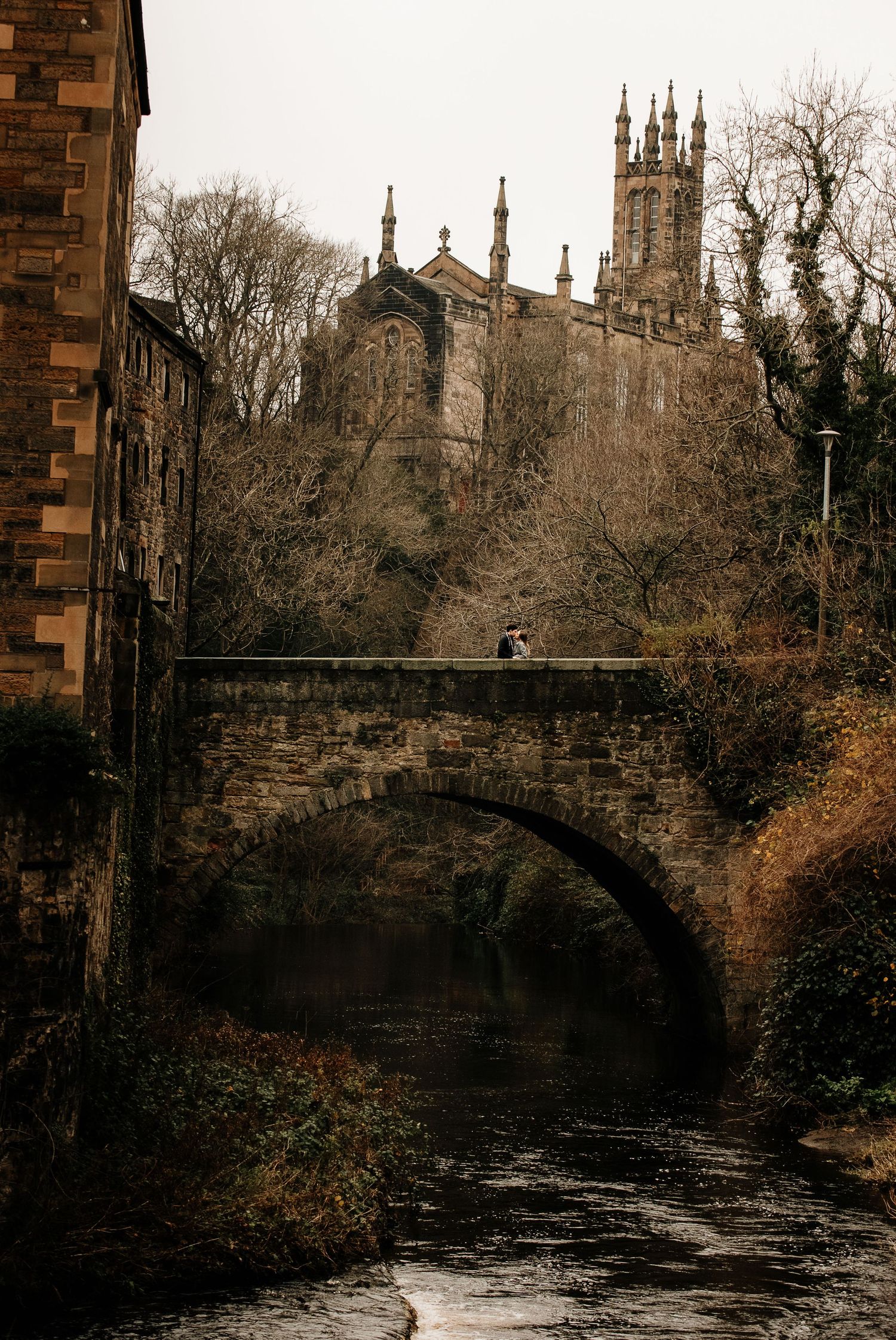 Ancient stone bridge spans across a river with gothic cathedral spires rising in the background.