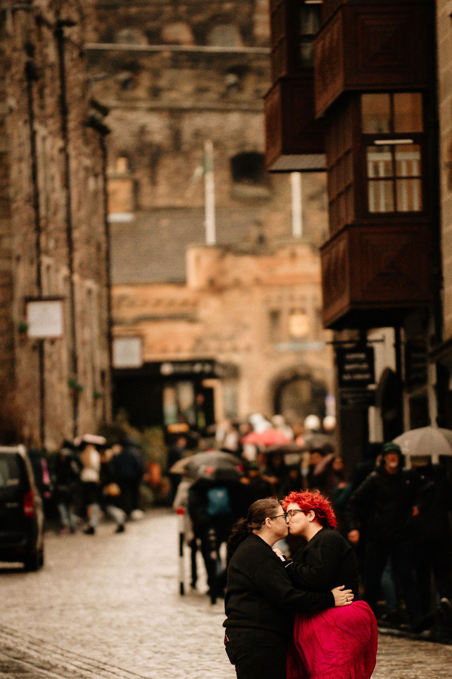 A romantic couple embraces in a red coat against a blurred urban European street backdrop with cobblestones.