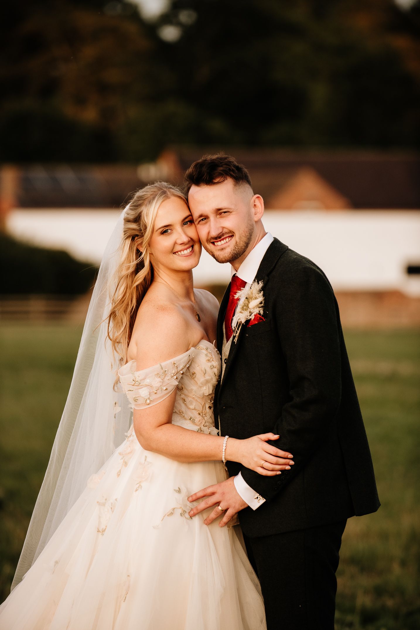 Smiling bride and groom pose outdoors in golden hour light, she in an off-shoulder gown with veil, he in a black suit.