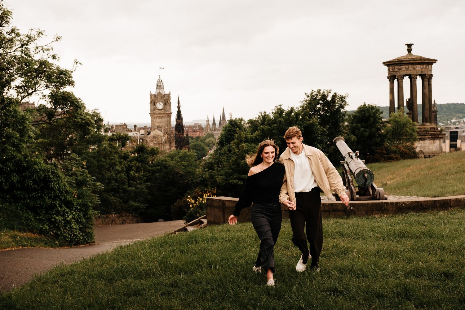 Couple walking hand in hand on Calton Hill, Edinburgh, with city skyline and Dugald Stewart Monument behind.